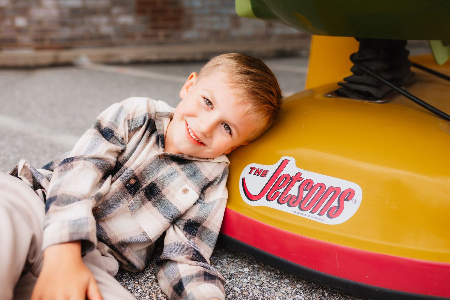 Smiling young boy in plaid shirt leaning against a yellow Jetsons ride-on toy outdoors.