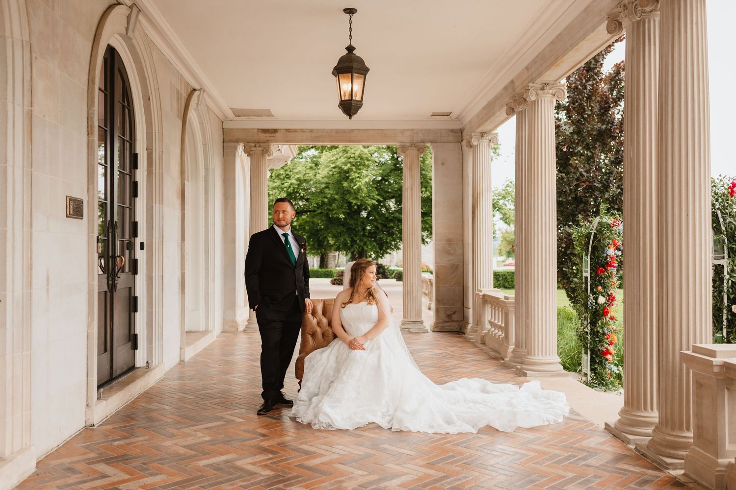 Bride in white gown seated on bench with groom on elegant columned porch of a classic mansion wedding venue.