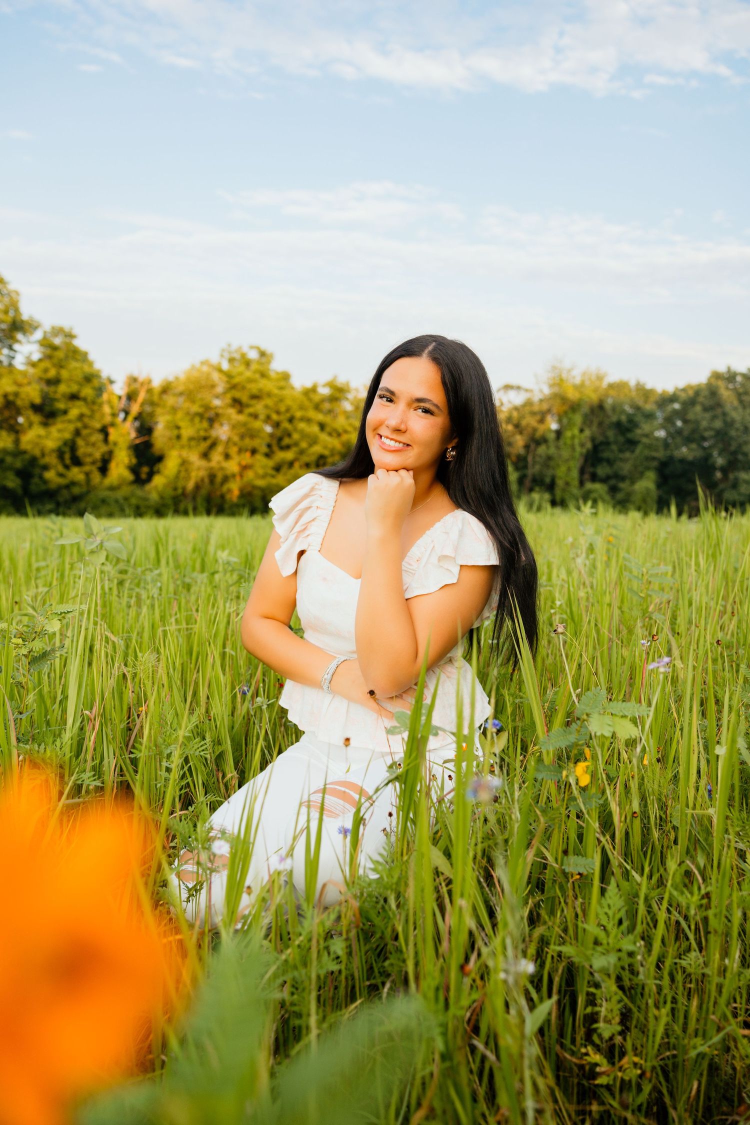 Smiling young woman in white dress sits peacefully among tall green grass and wildflowers at golden hour.