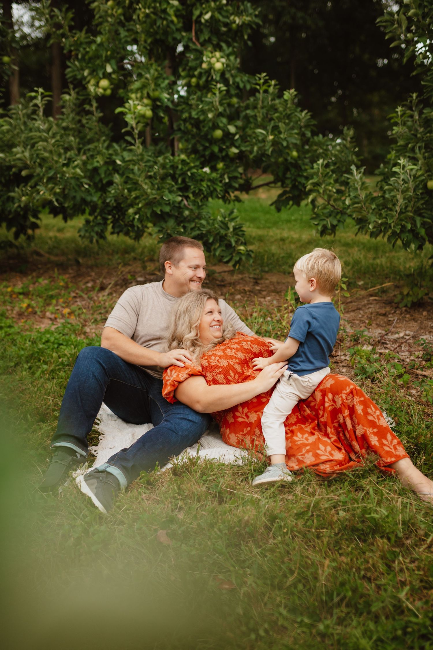 A smiling family of three relaxes together on a blanket in an apple orchard, with a toddler playfully leaning on his parents in the grass.