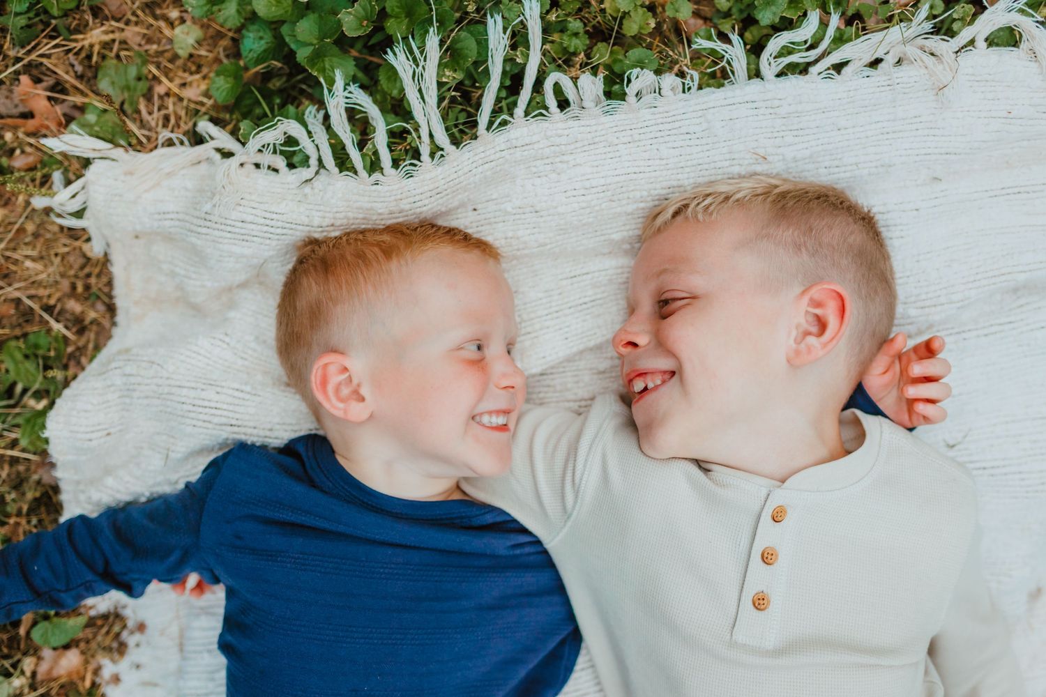 Two smiling young boys lie side by side on a white fringed blanket over green grass and leaves, laughing and looking at each other.