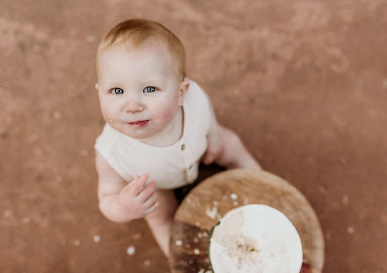 Adorable baby in white onesie smashing first birthday cake on terracotta floor looking up at camera.
