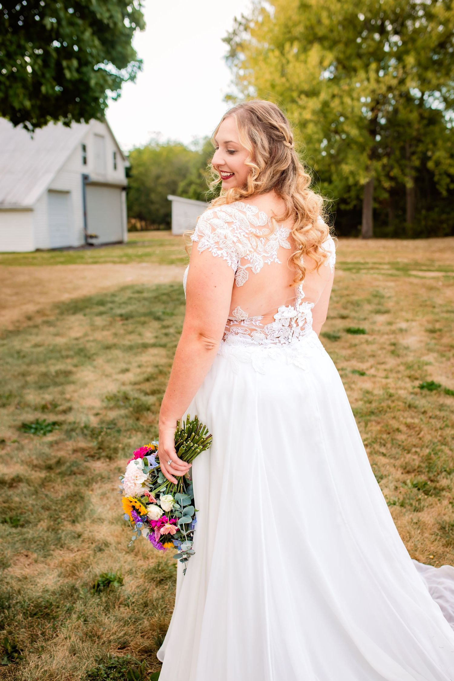Smiling bride in lace back wedding gown holding colorful bouquet in outdoor setting.