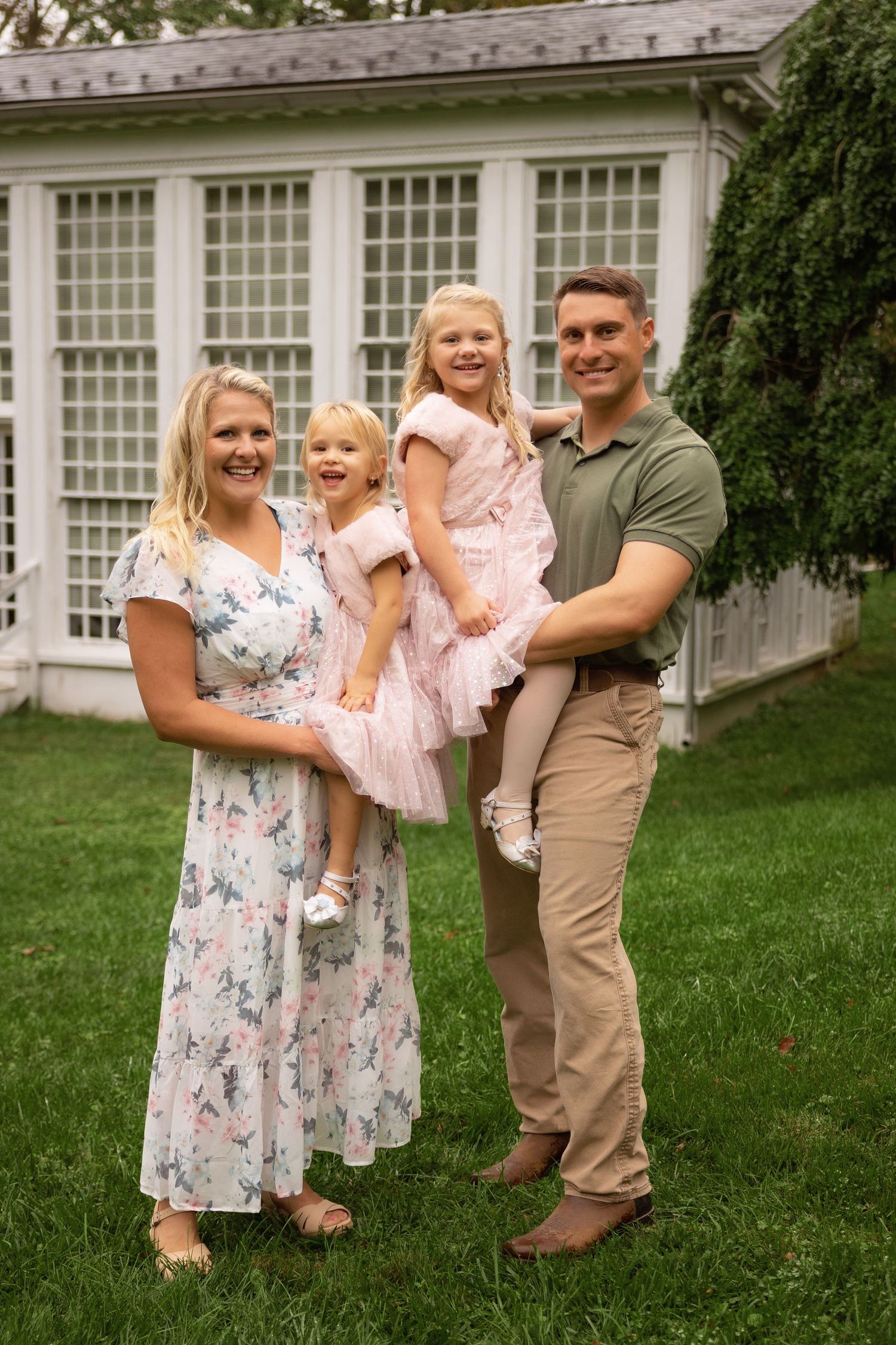 A smiling family of four poses on a green lawn, with a mom in a floral dress, a dad in a green polo, and two girls in pink dresses.