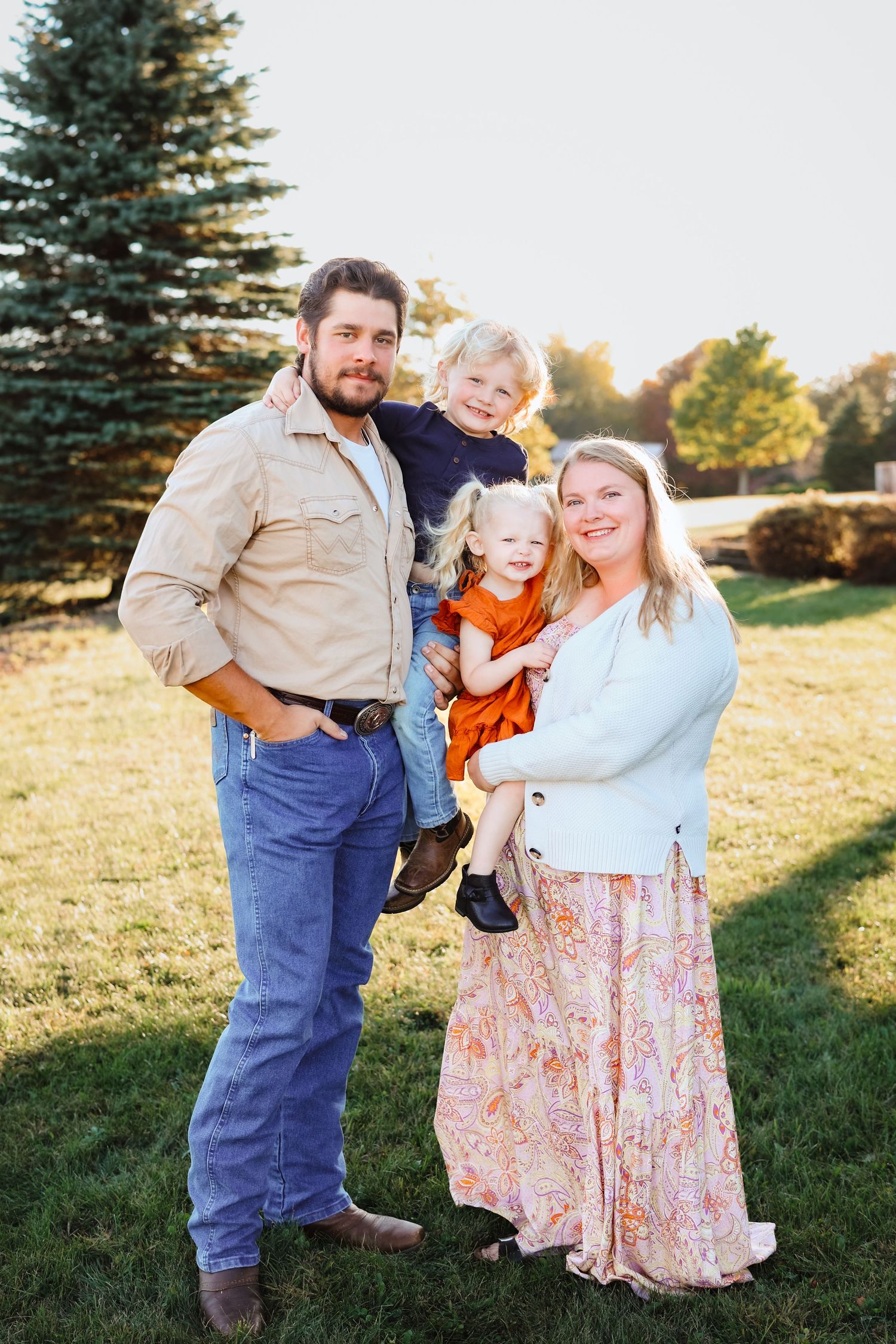 Young family of four with two toddlers pose together on a sunny green lawn outdoors.