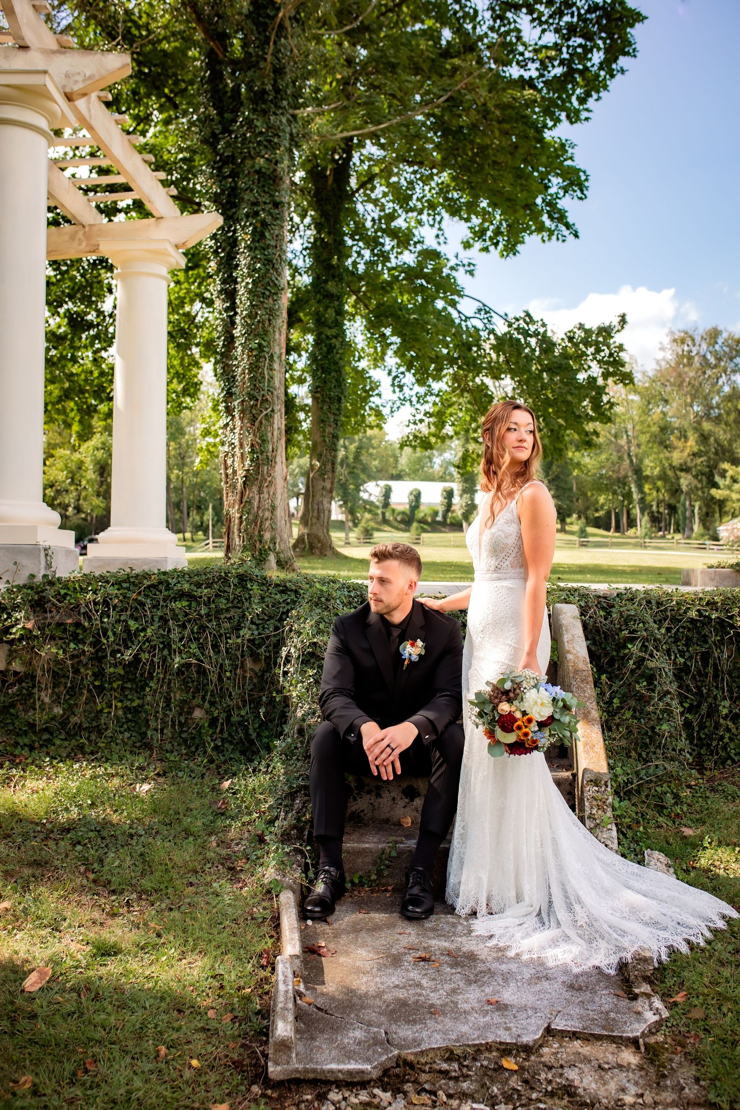 Bride and groom pose near ivy-covered white pergola on stone steps in a sunny garden.