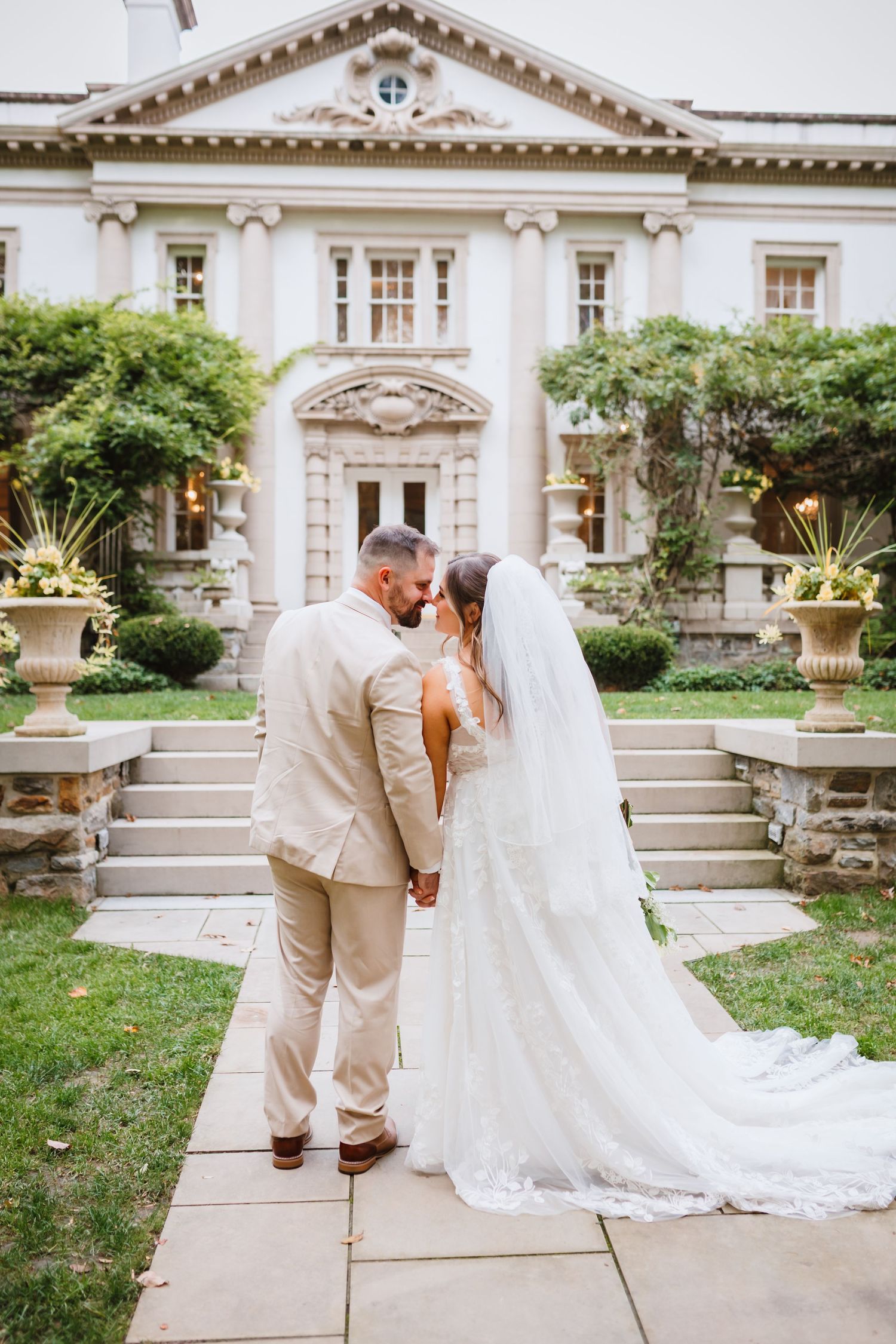 Bride and groom kiss on a stone pathway in front of an elegant white mansion wedding venue.