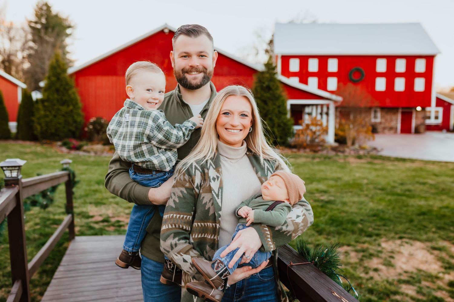 Smiling family of four posing on wooden bridge with charming red barn in background.