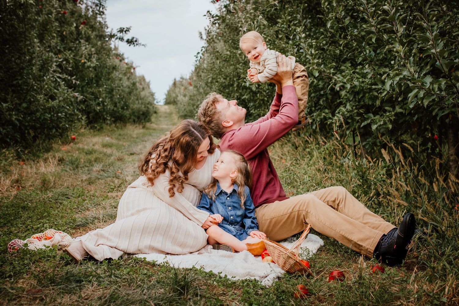 Young family of four sitting on a blanket in an orchard, lifting baby joyfully.