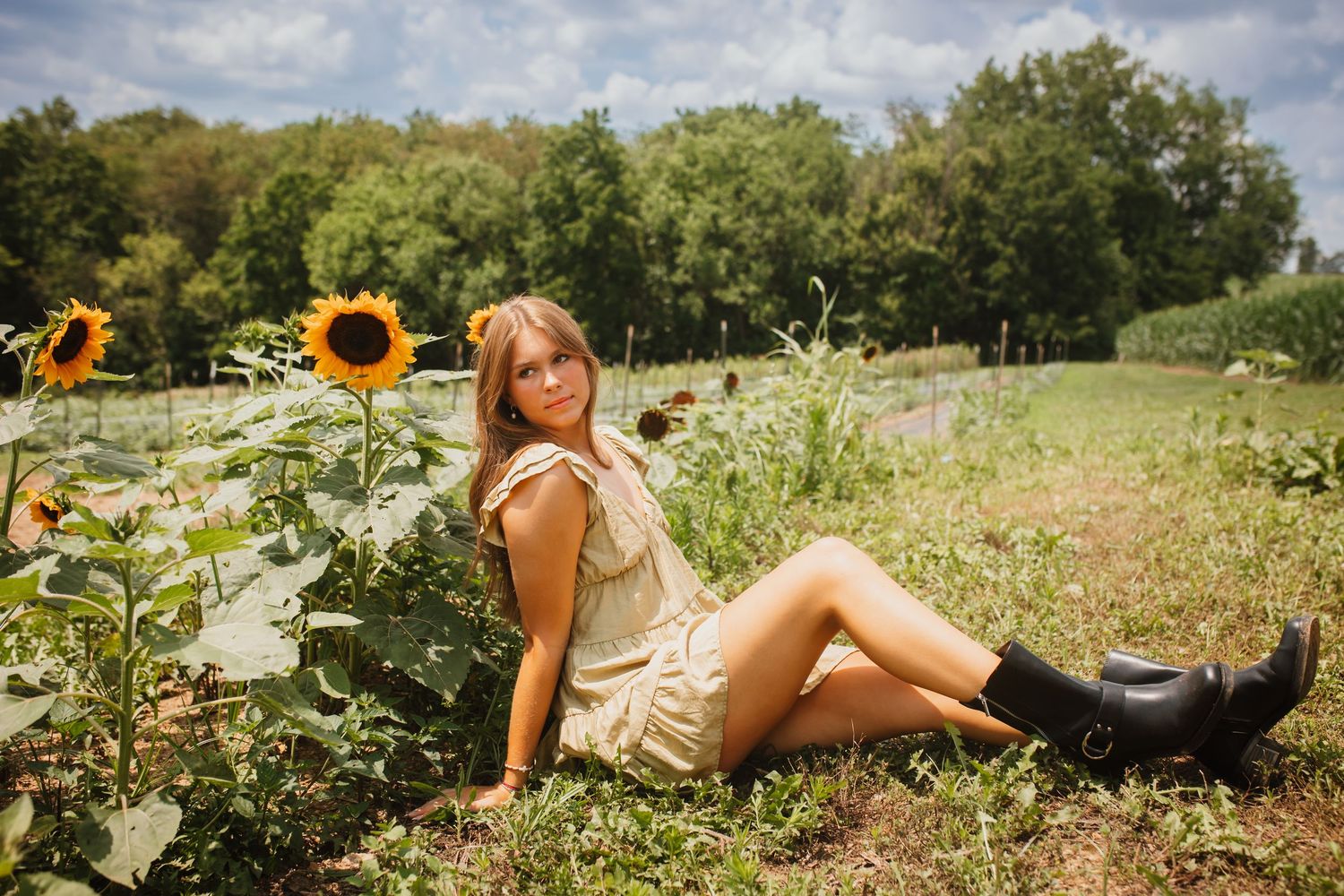 Young woman in a cream dress and black boots sitting beside sunflowers in a field.