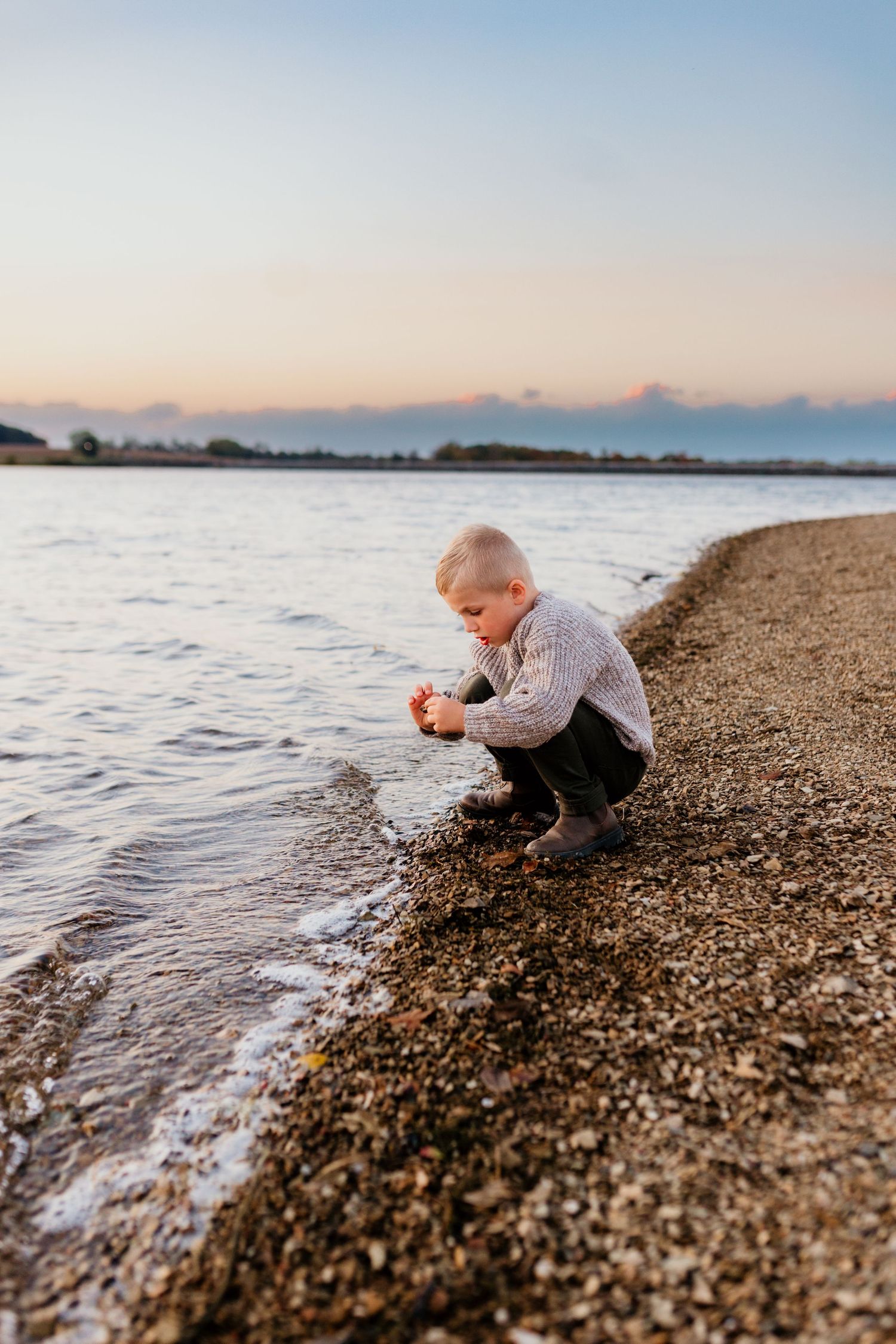 Young boy crouching on a rocky shoreline exploring the water at dusk.