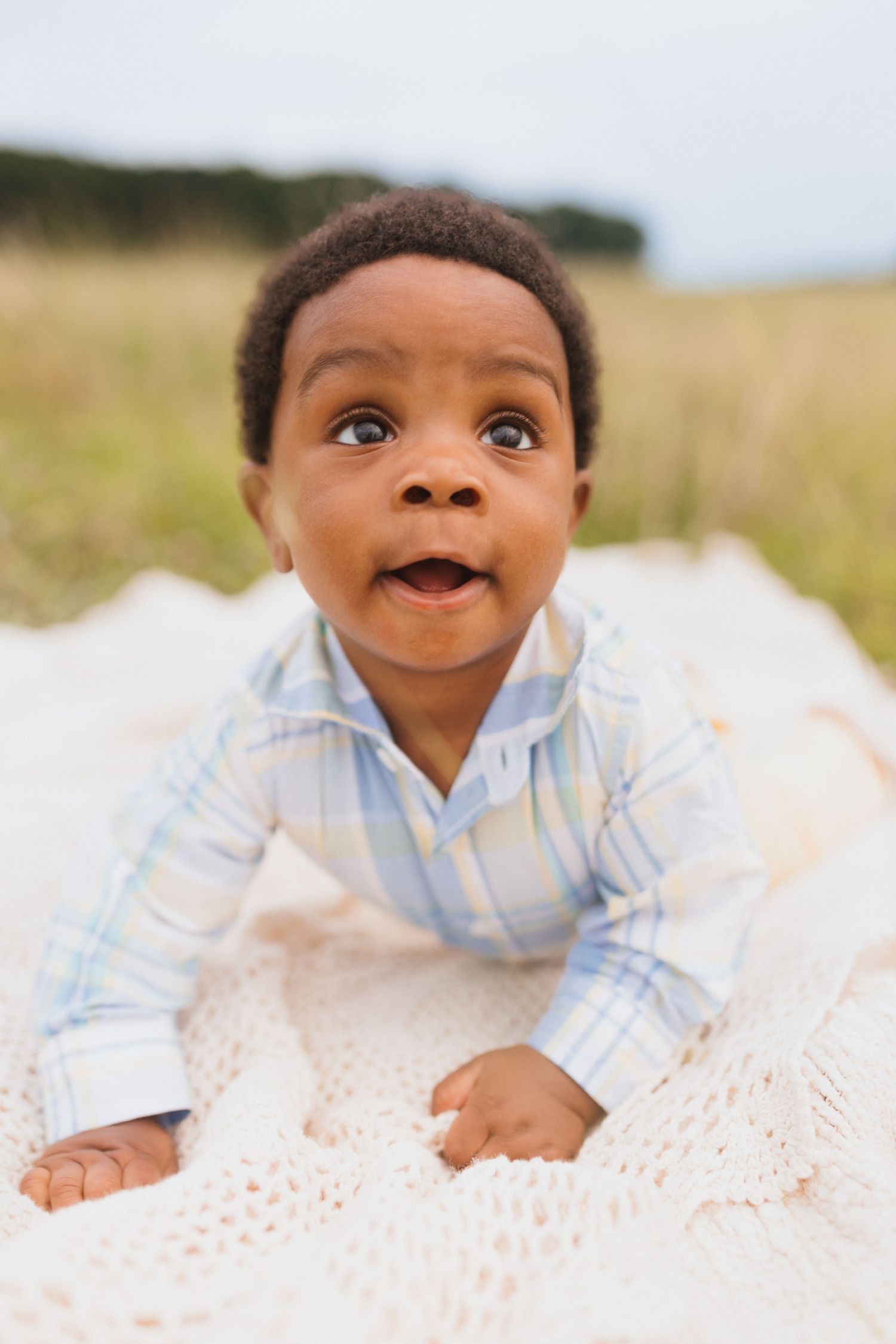 Adorable Black baby in plaid shirt doing tummy time on white blanket in field.