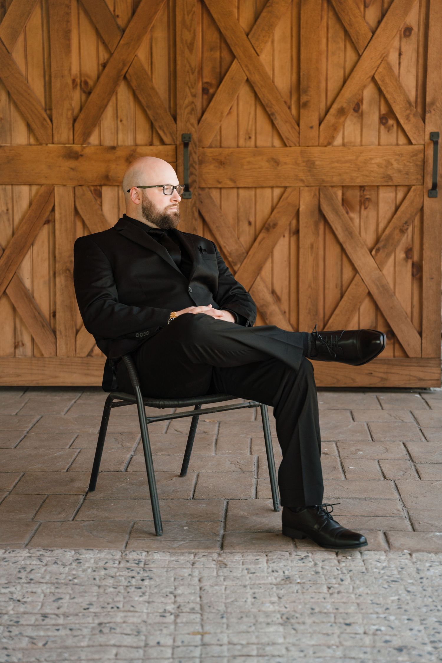 Bald bearded man in black suit sitting on chair with legs crossed in front of wooden barn doors.