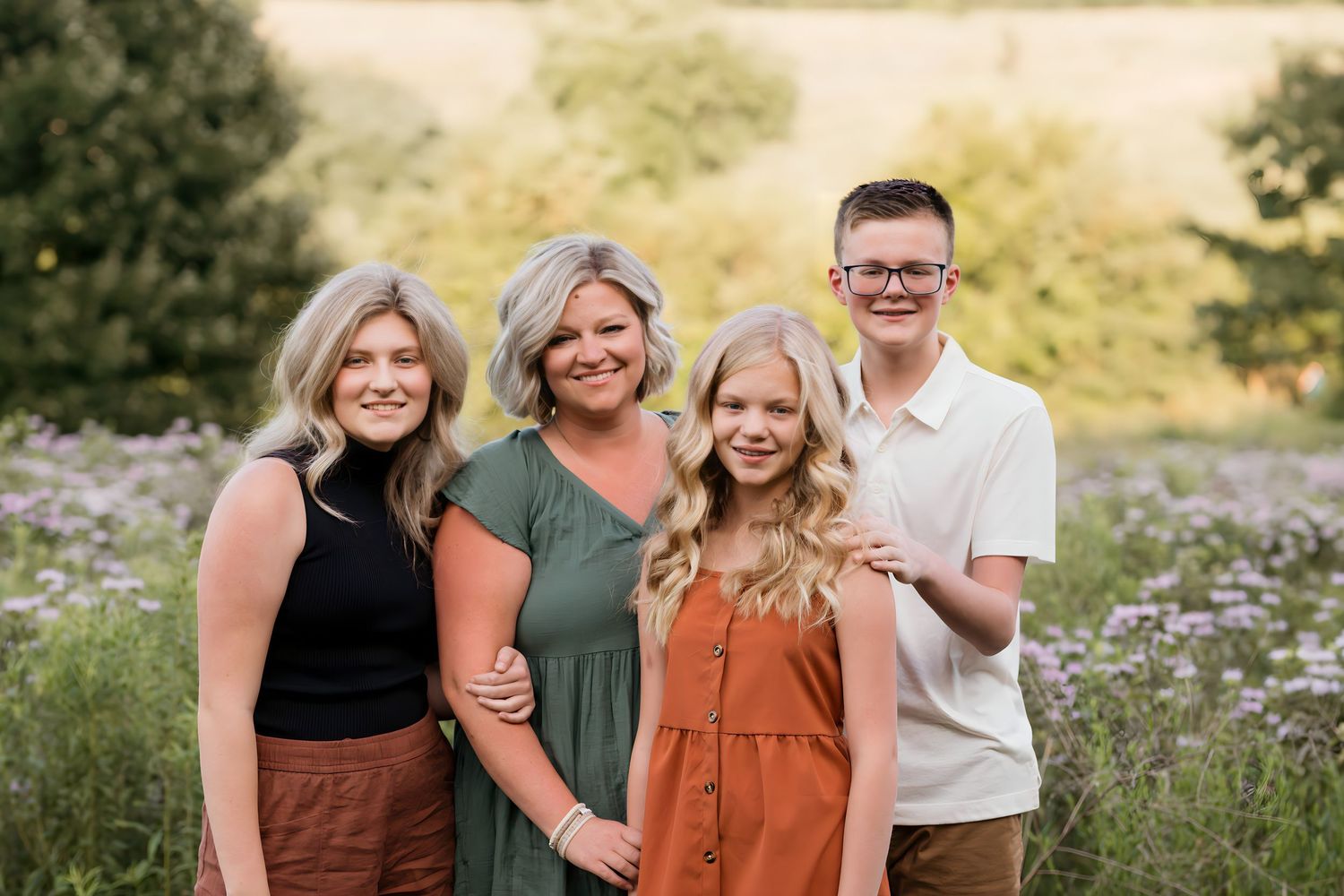 A family of four smiles together in a field of purple wildflowers at sunset.
