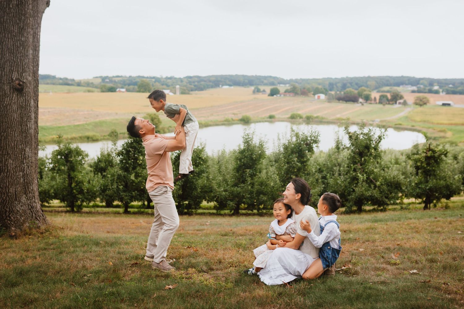 Family of five enjoying an outdoor photo session on a grassy hillside, with a father lifting a child overhead near a scenic lake and trees.