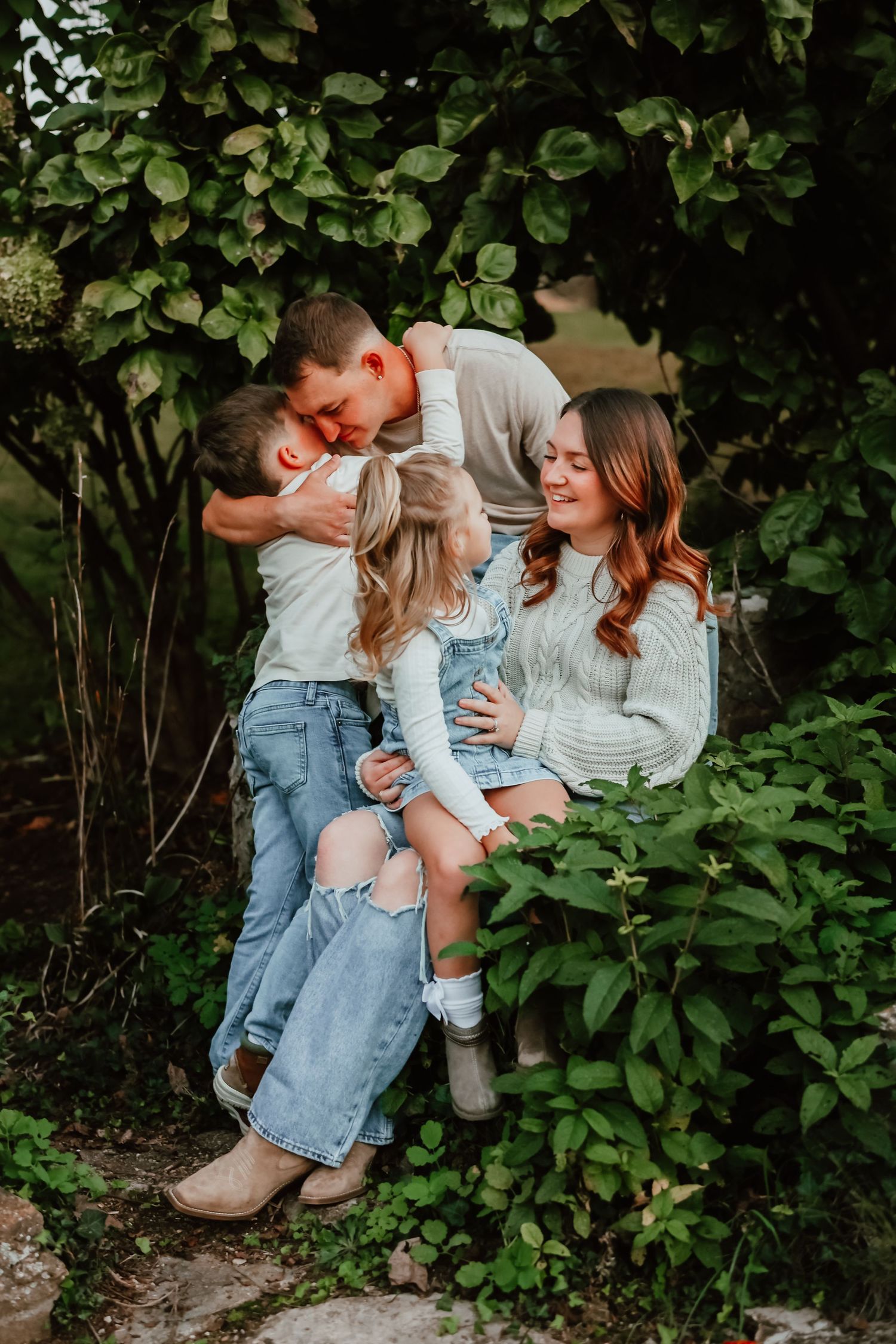 Affectionate family of four cuddling together beneath lush green garden foliage.