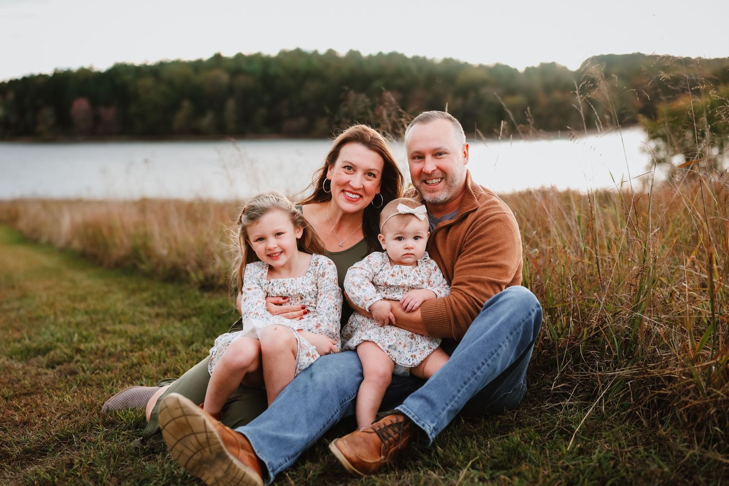 Smiling family of four with two young daughters sitting by a peaceful lakeside.