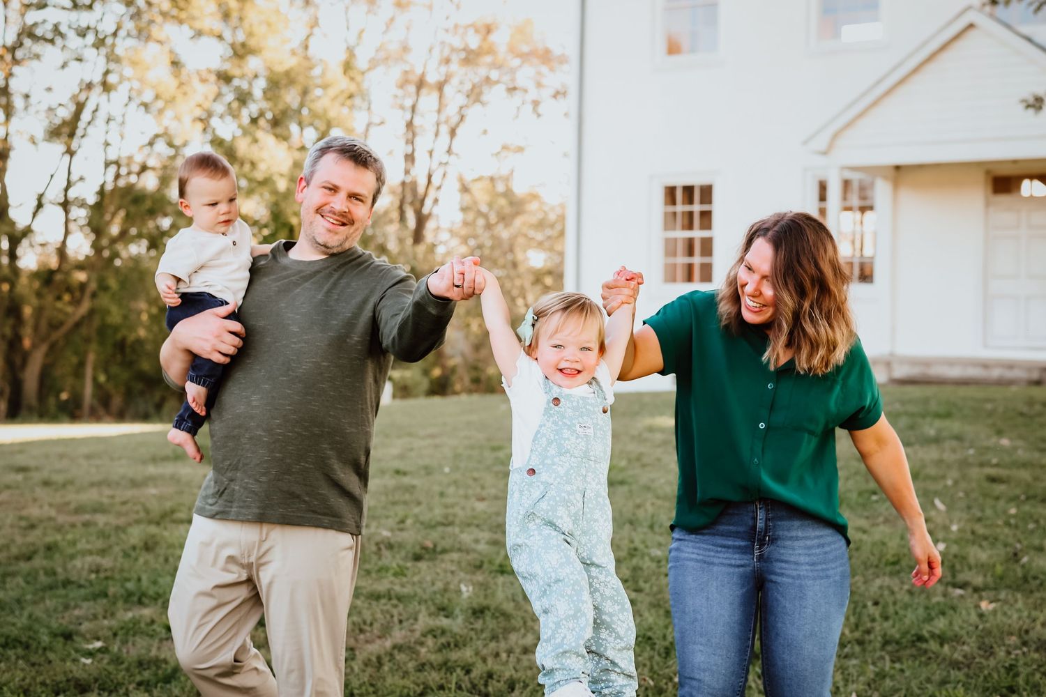 Joyful family of four plays outdoors on green grass in front of a white farmhouse.