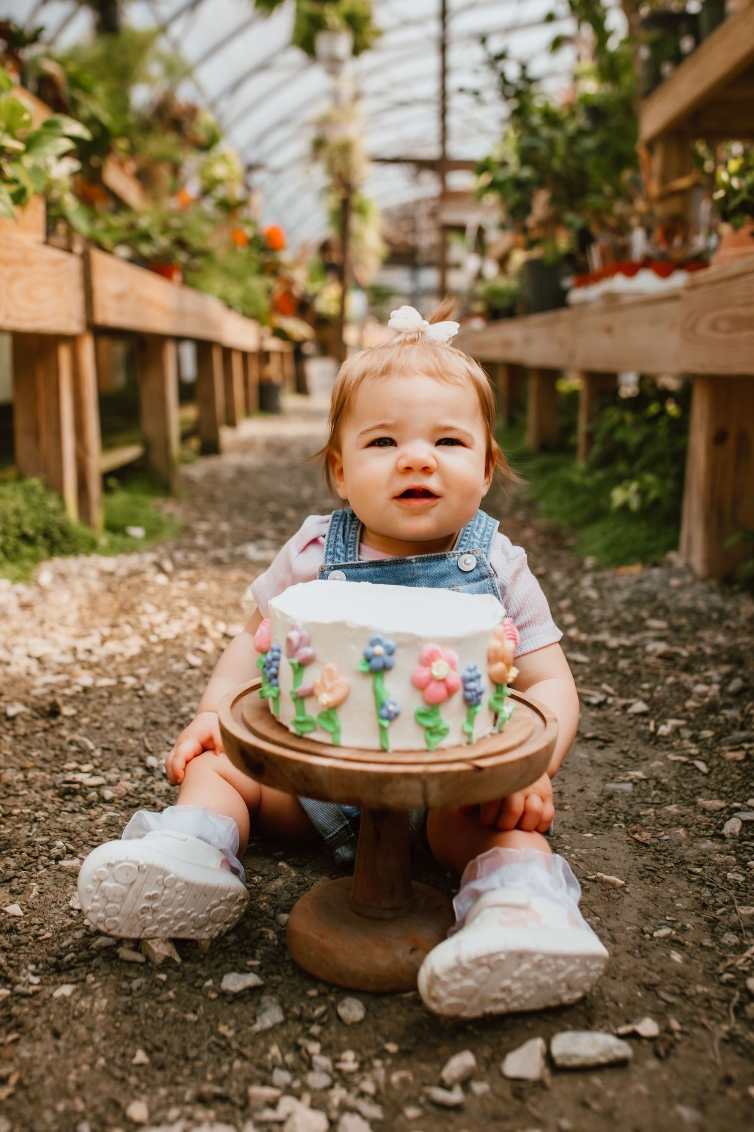 Adorable baby girl in denim overalls holding a floral decorated white birthday cake in a greenhouse.