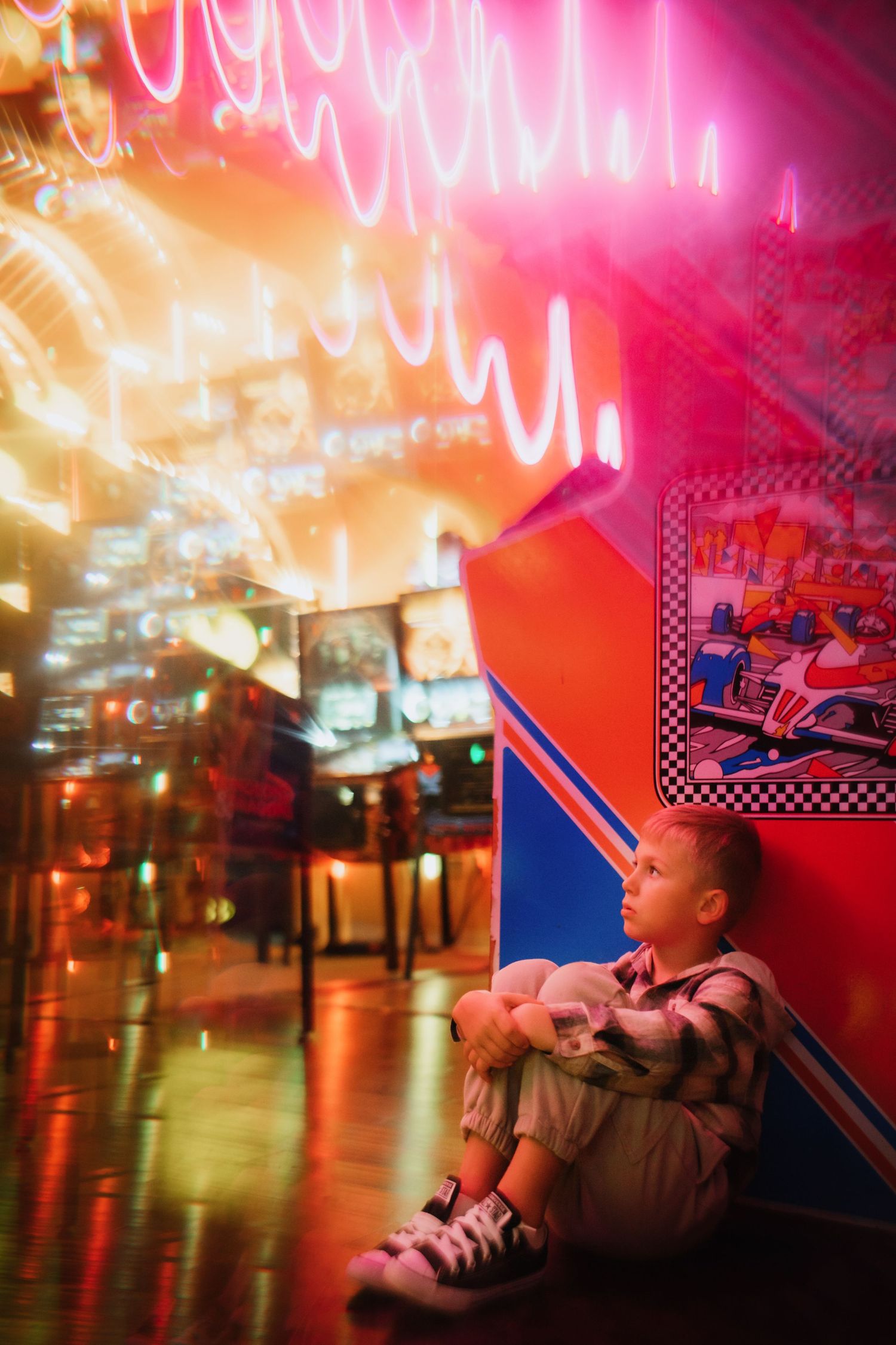 Young boy sitting on floor hugging stuffed animal beside glowing neon arcade game machines.
