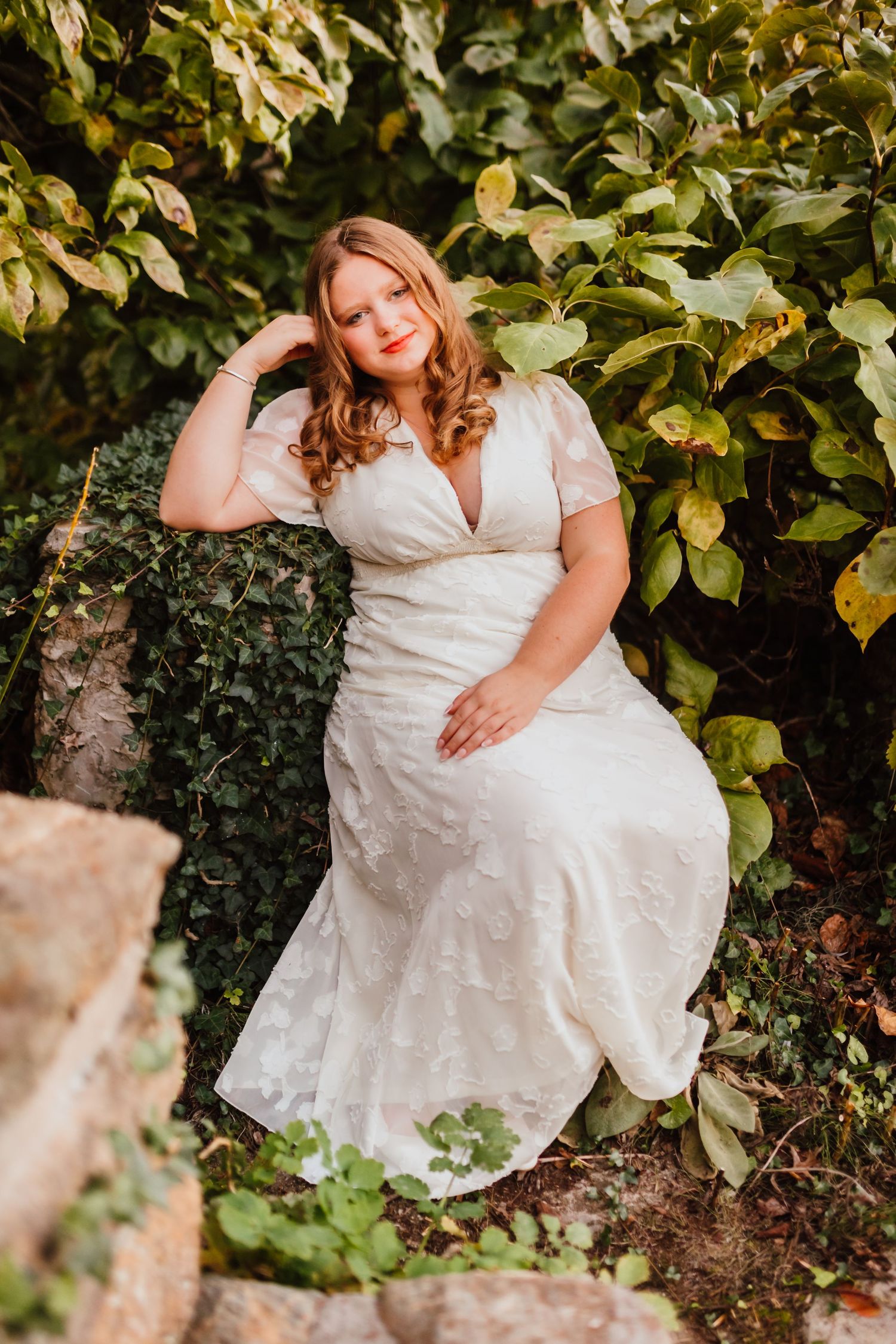 Plus-size bride in white lace wedding gown poses among lush green garden foliage.