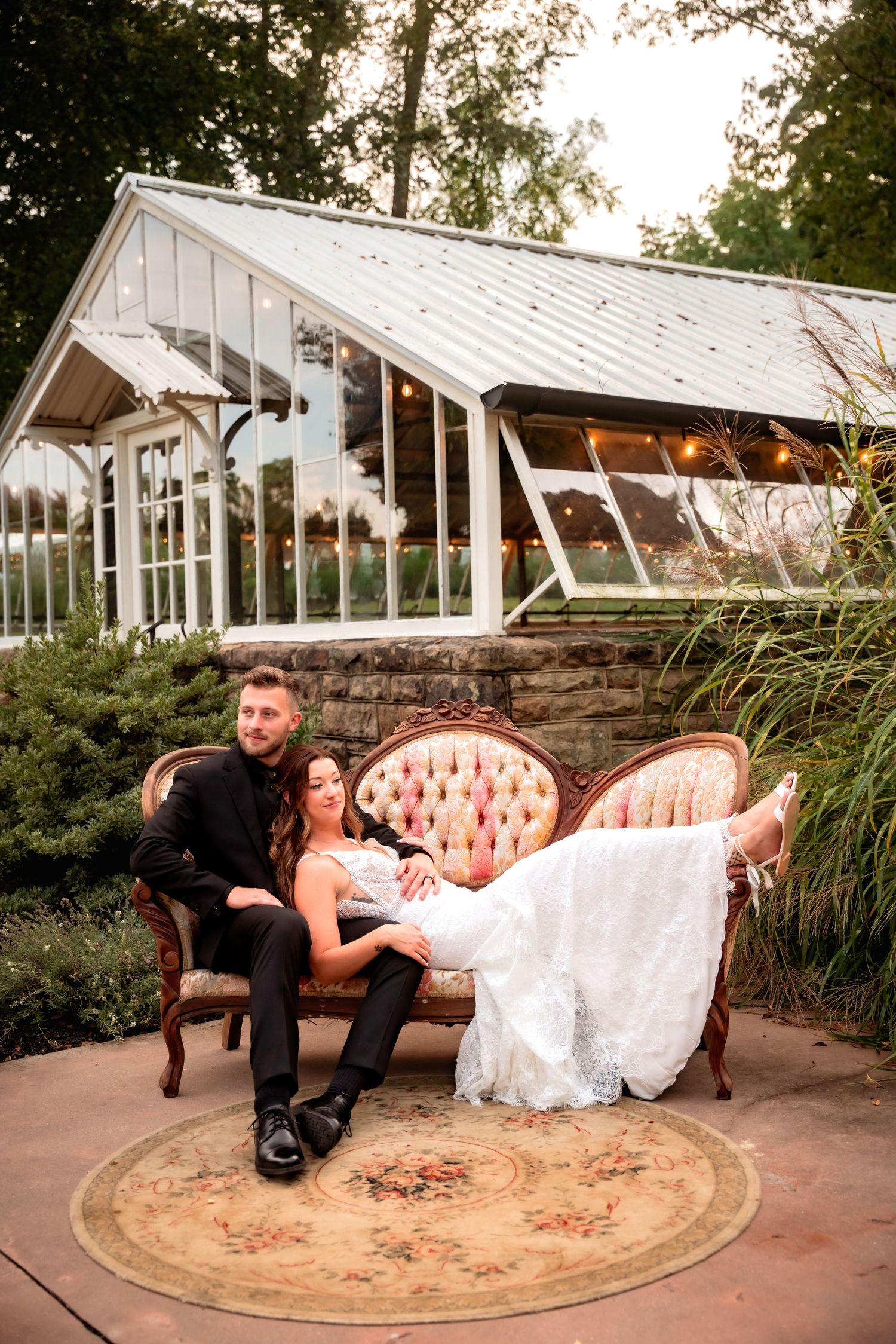 Bride and groom relax on a vintage pink sofa in front of a glass greenhouse at dusk.