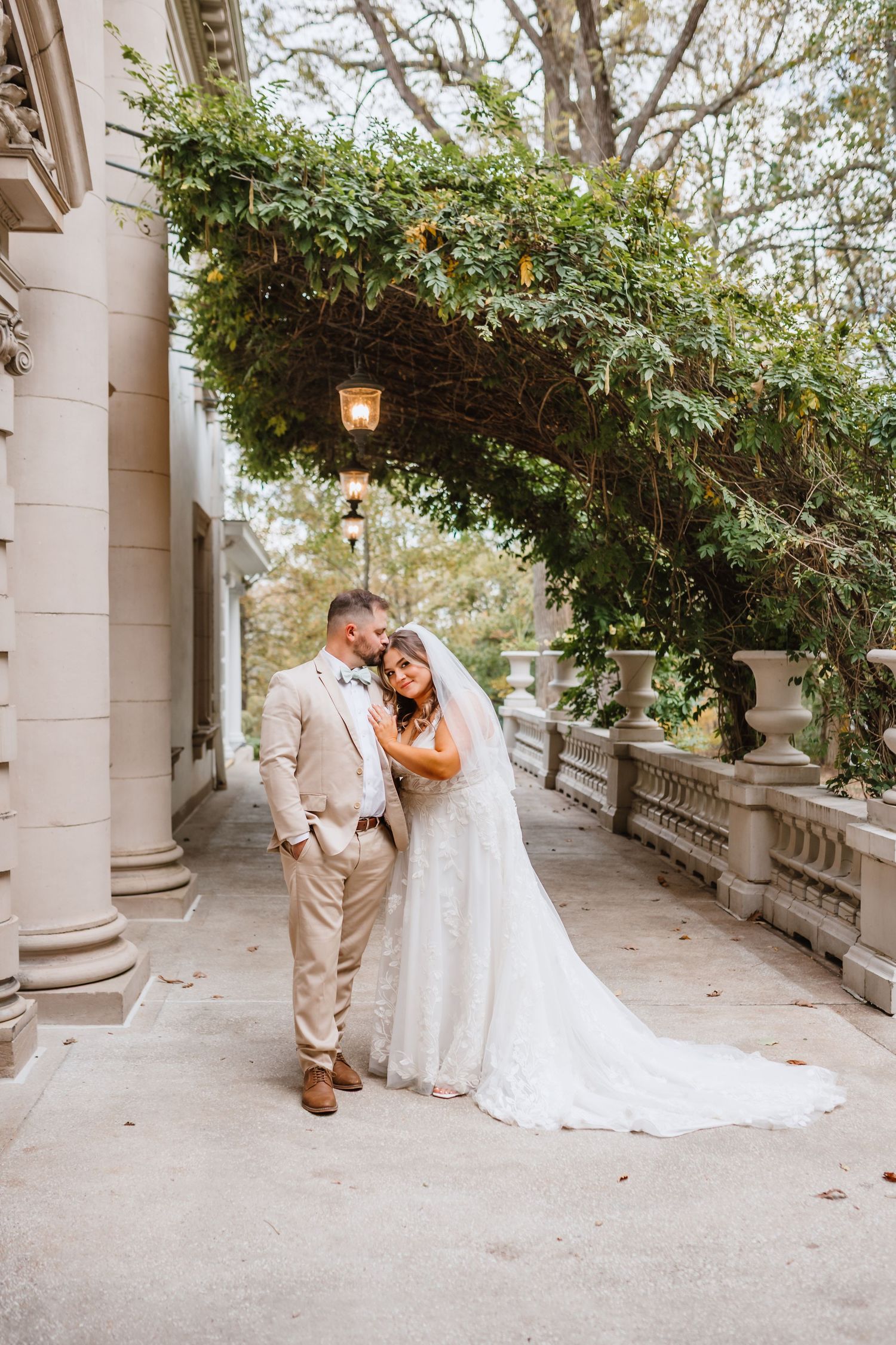 Bride and groom share an intimate moment under a lush green archway on a grand estate balcony.