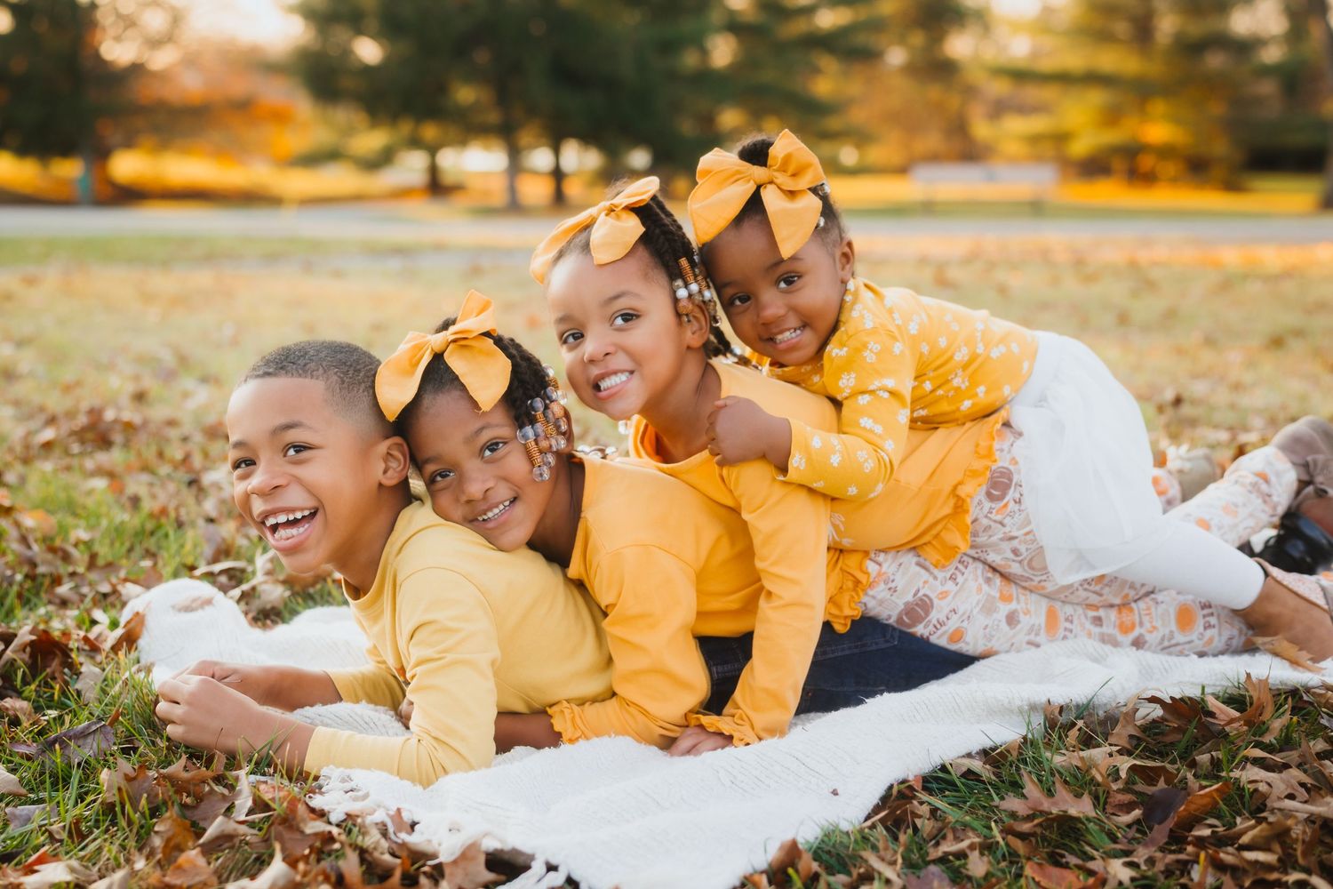 Four Black children in matching yellow outfits lying in autumn leaves on white blanket during fall photo session.