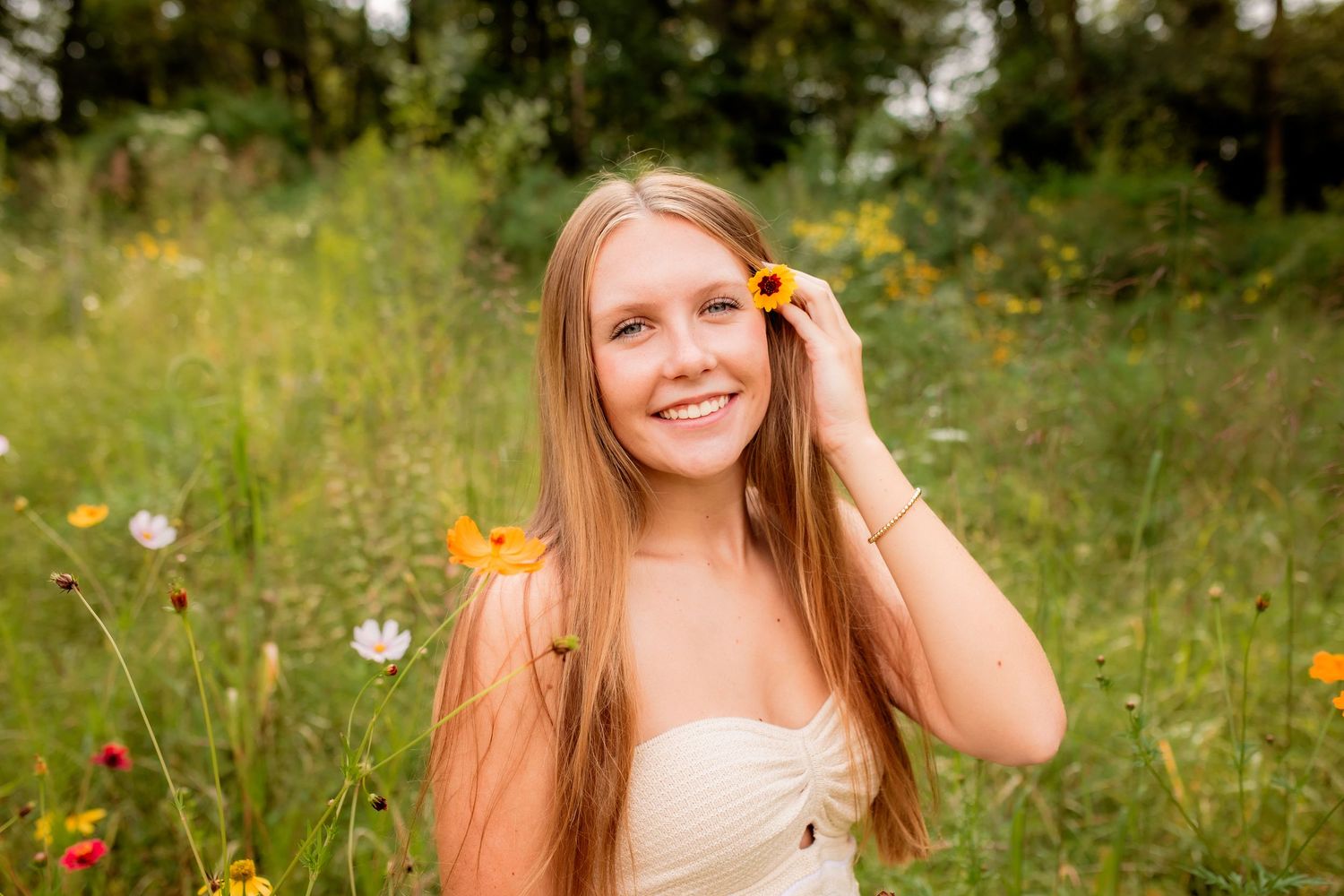 Smiling blonde young woman holding a sunflower in a wildflower meadow for a senior portrait session.