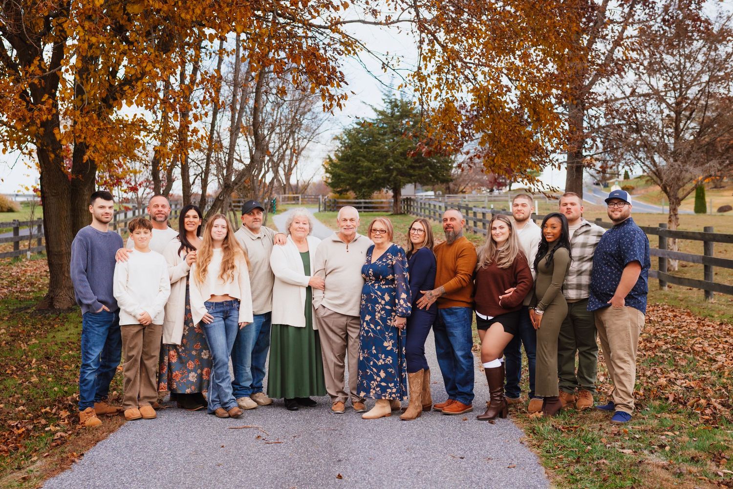 Large multigenerational family of thirteen posing on rural driveway surrounded by colorful fall foliage.