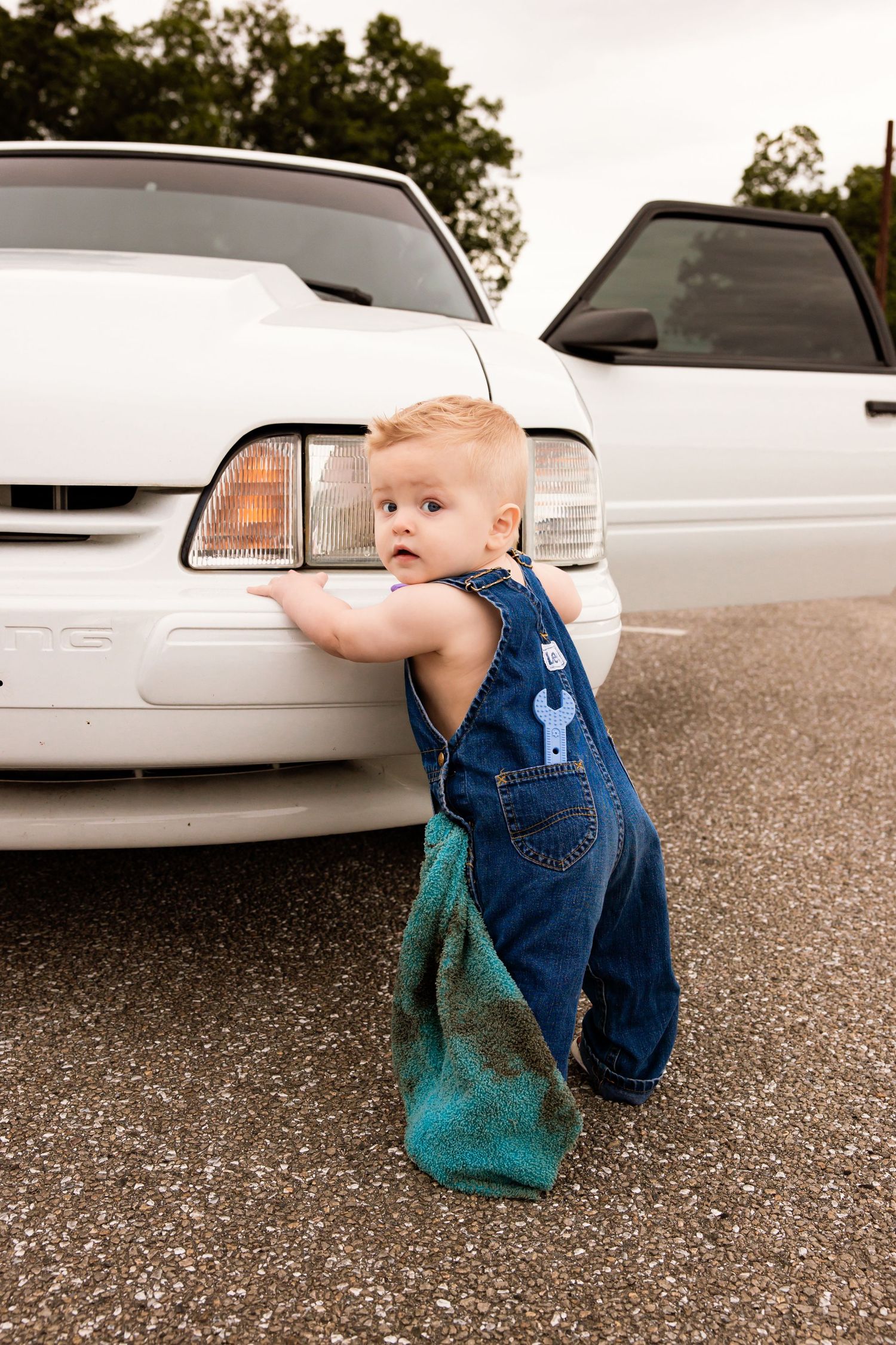 Toddler in denim overalls standing next to a white car in a parking lot.