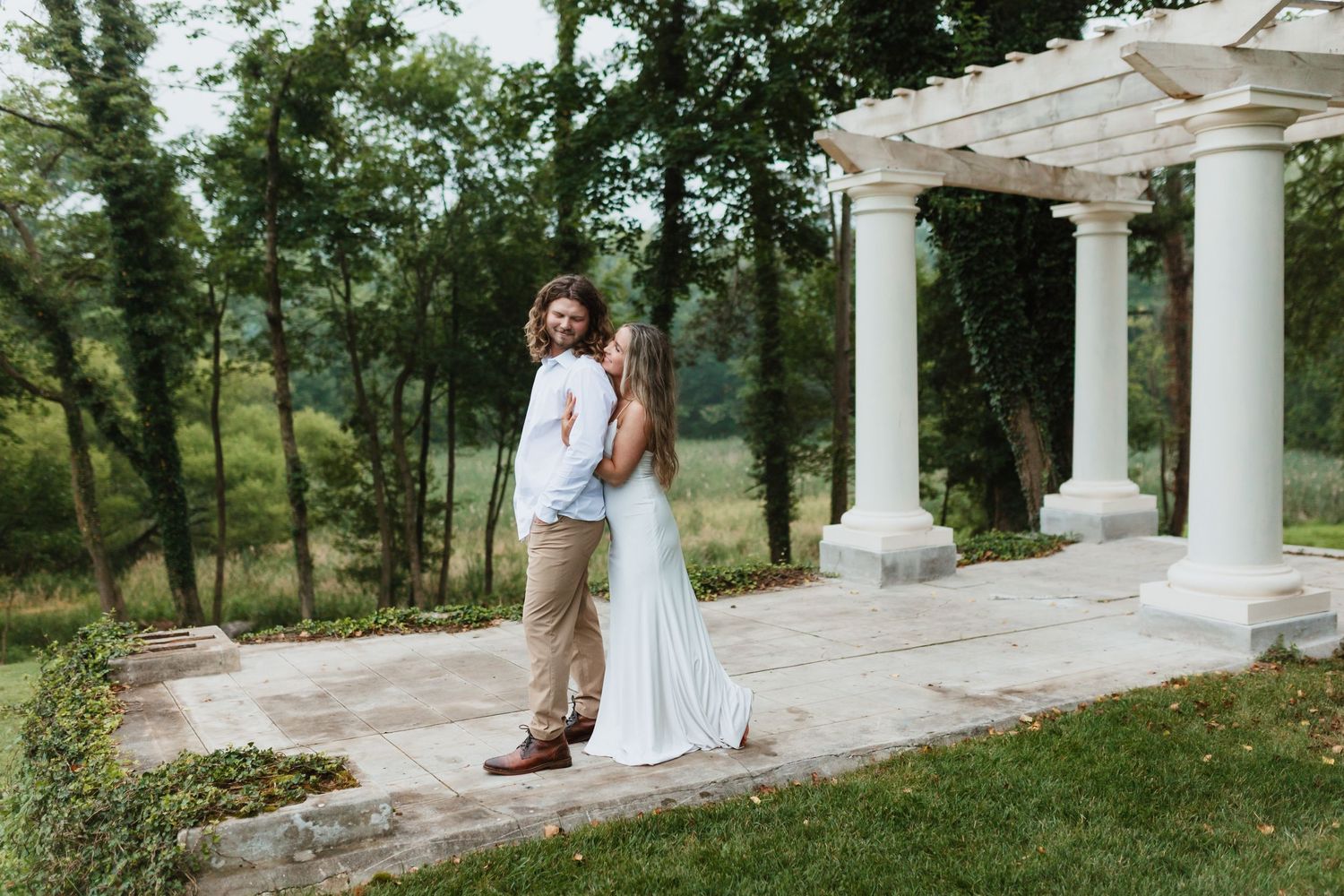 Romantic couple dancing near a white pergola surrounded by lush green trees in a garden setting.