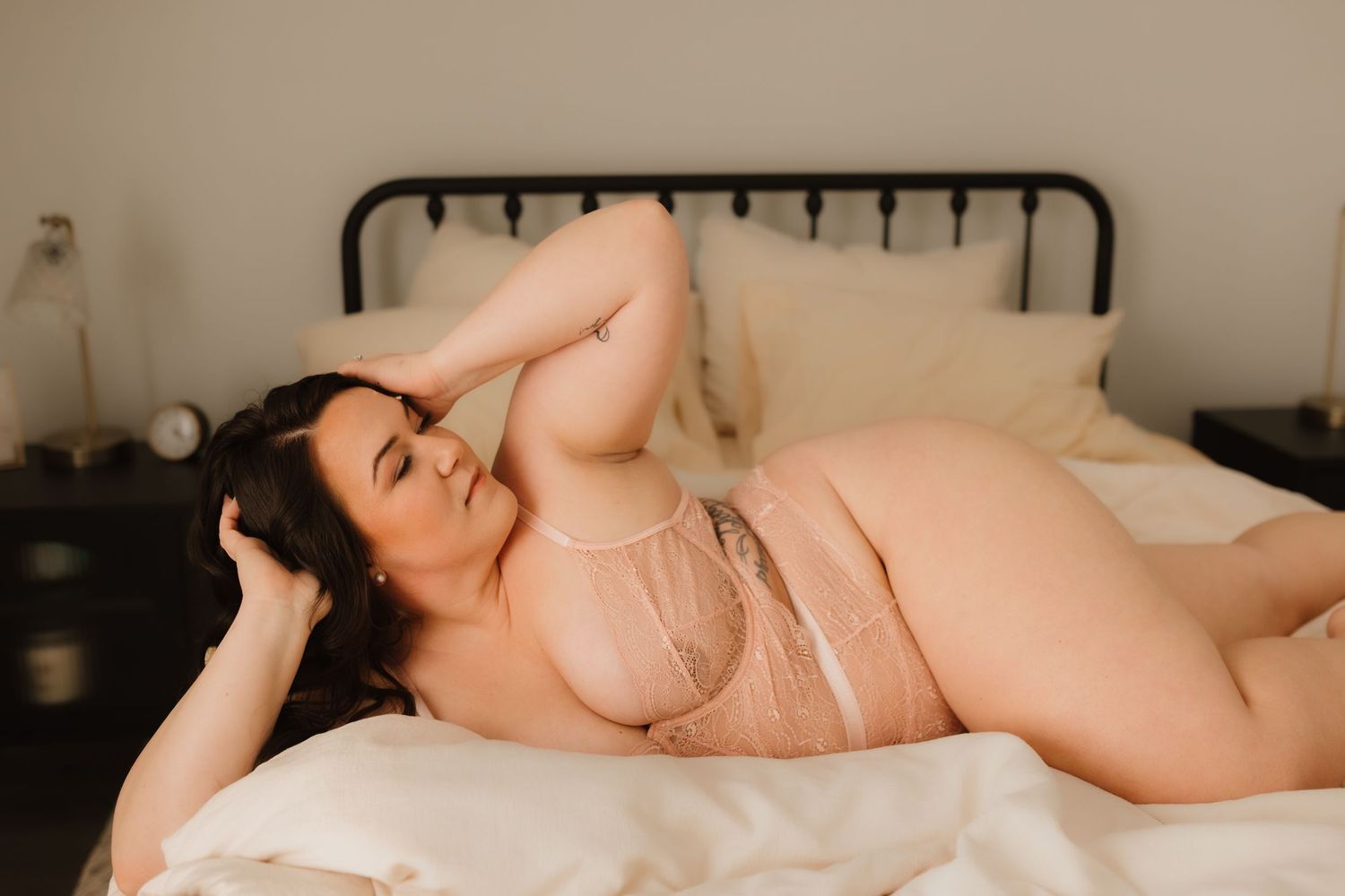 Brunette woman in sheer pink lingerie lying on white bed in soft light.