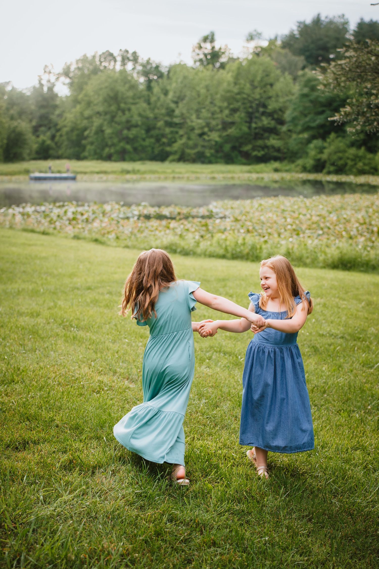Two young girls in teal and blue dresses spinning and holding hands on lush green lawn by pond.