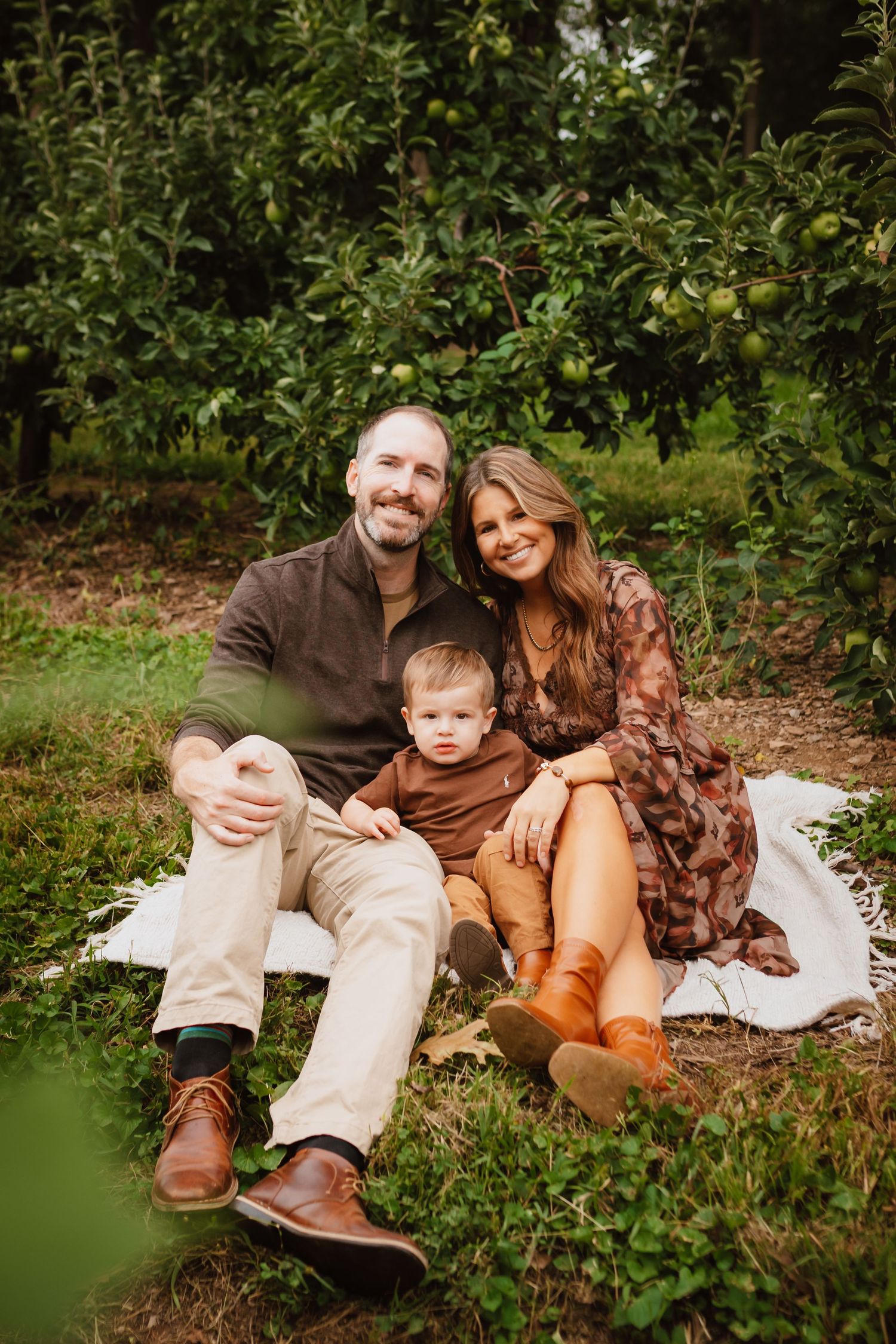 Smiling couple sitting on a white blanket with their toddler in an apple orchard, wearing cozy fall outfits and brown boots.