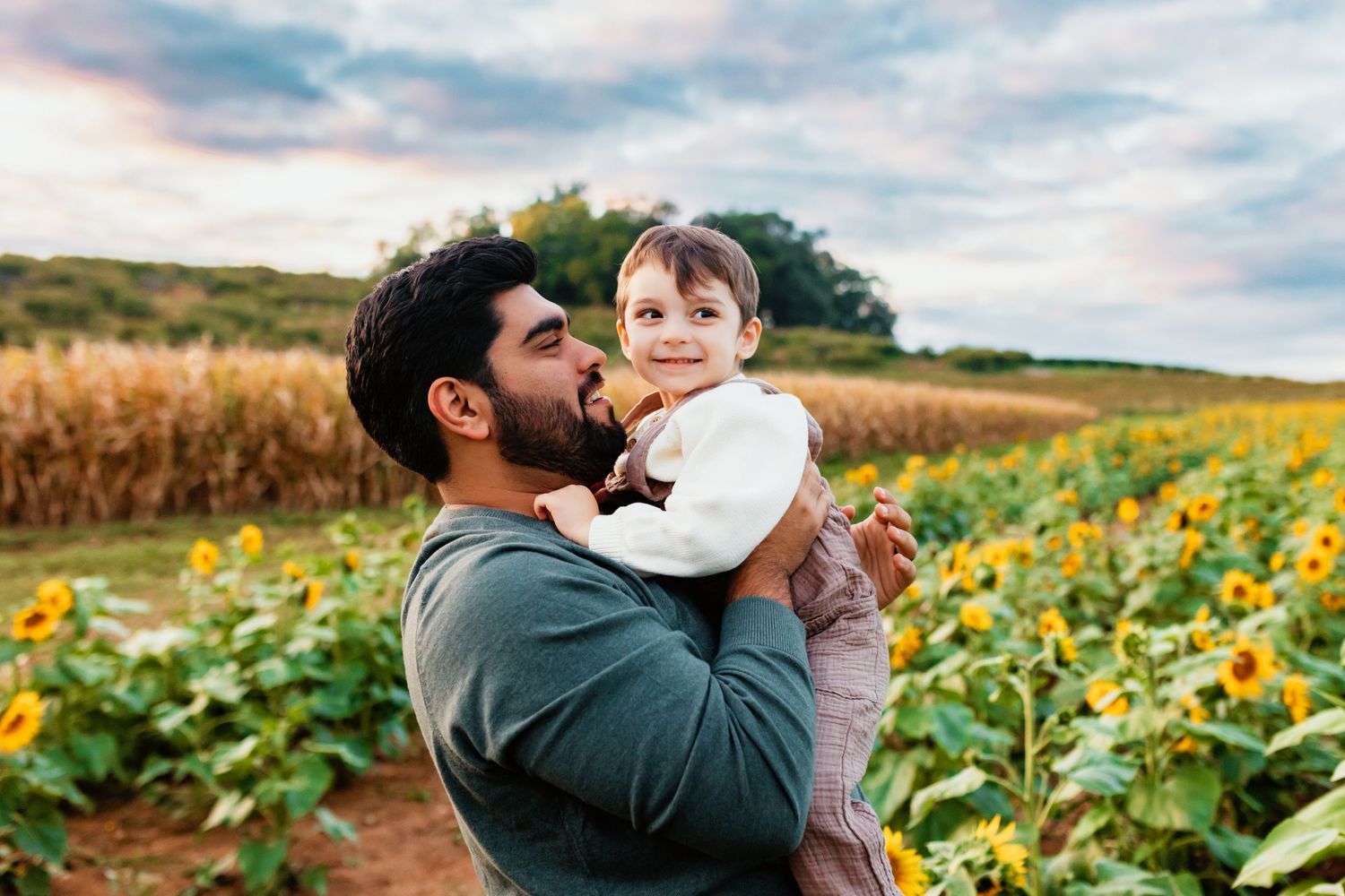 Father holding smiling toddler in sunflower field with corn crops in background at sunset.