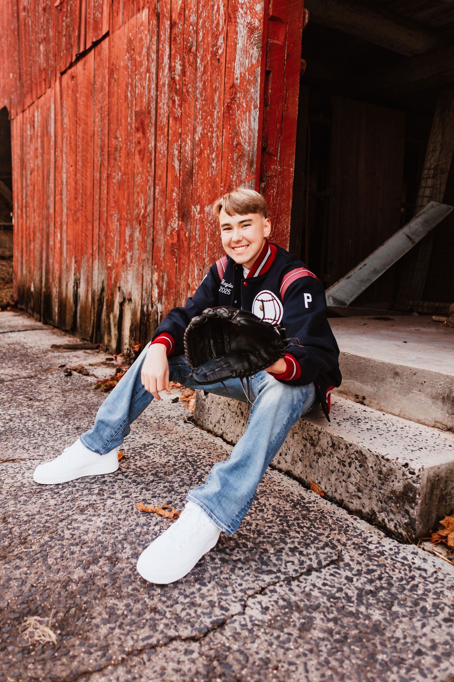 Smiling teen in varsity jacket holding baseball glove seated on steps by rustic red barn.