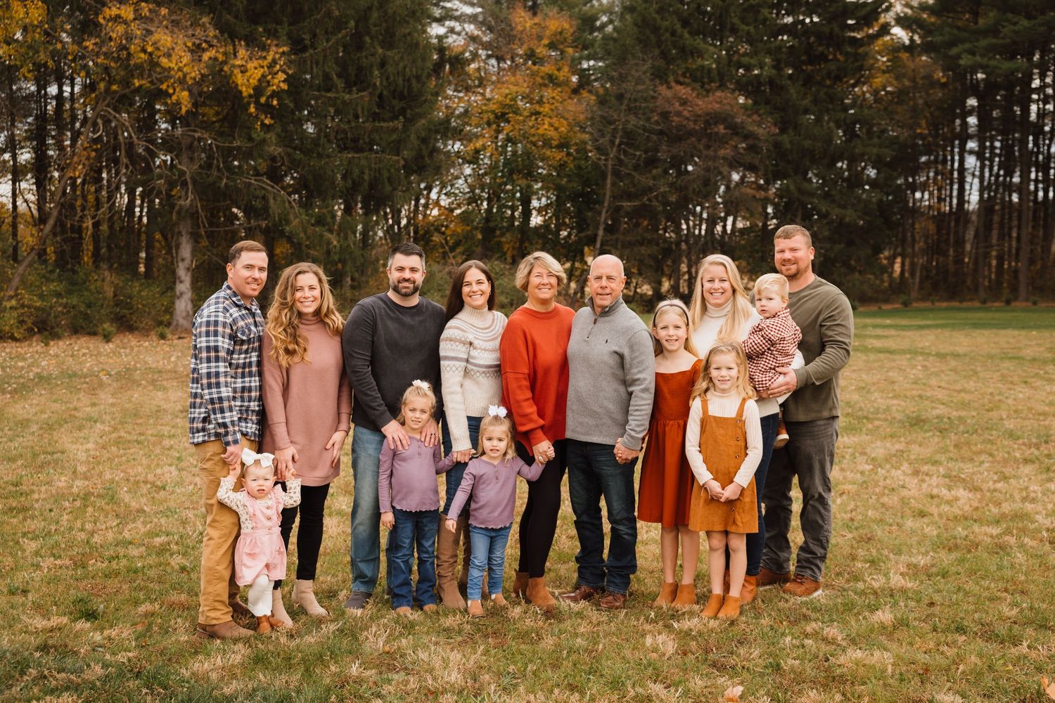 Large extended family posing together in a fall field surrounded by colorful trees.