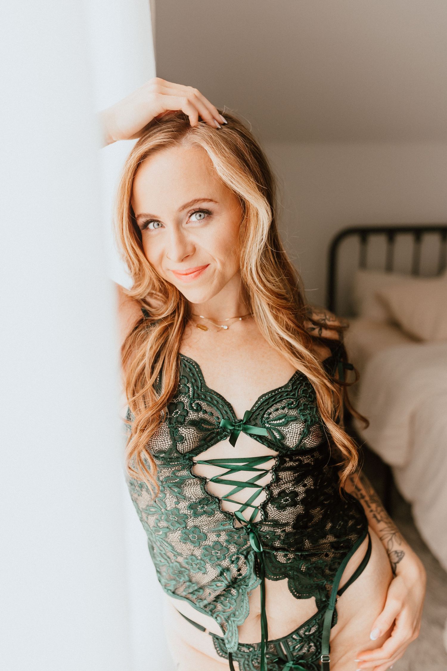 Blonde woman in green lace corset lingerie posing by a white wall in a bright bedroom.