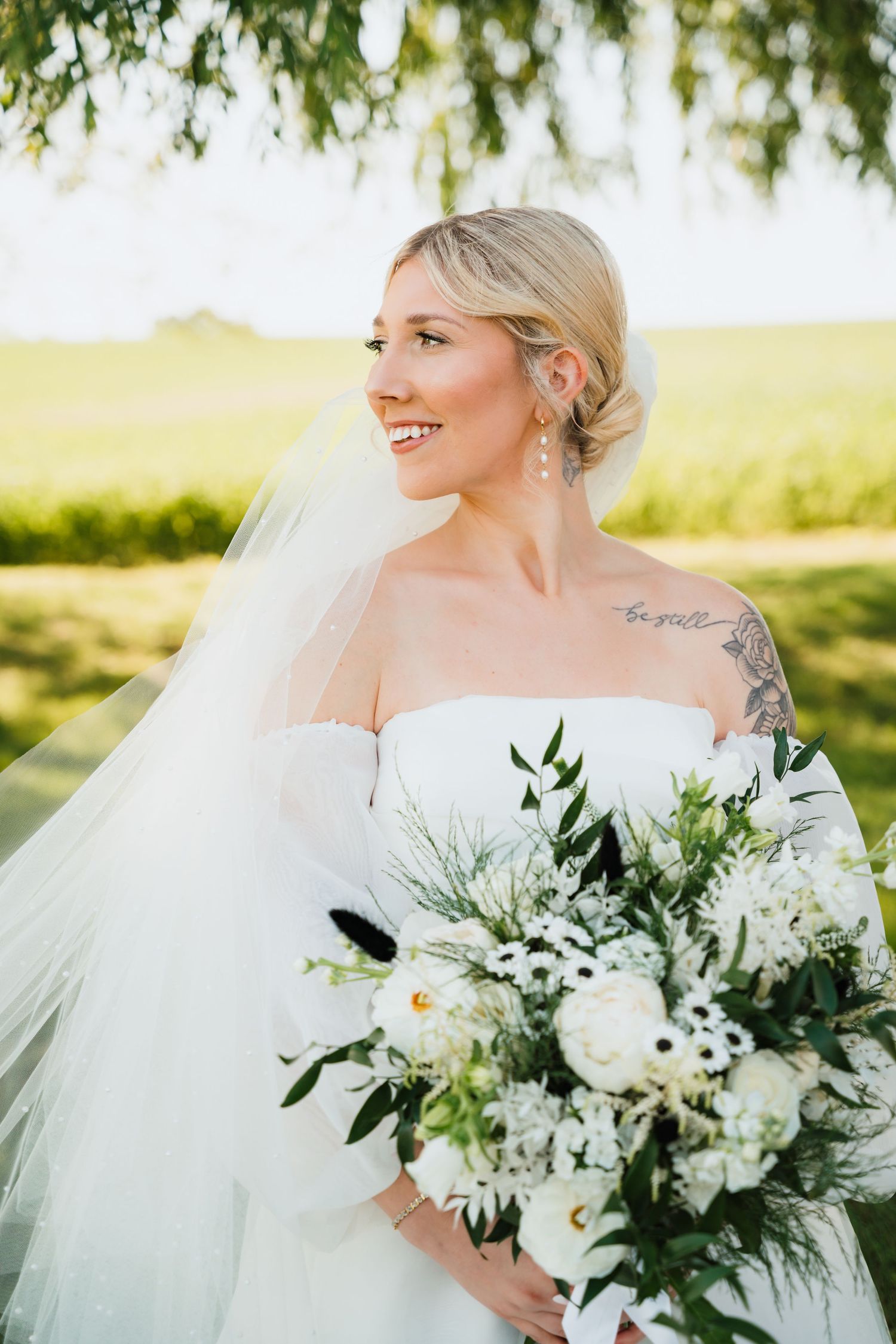 Radiant bride in strapless gown with veil and floral bouquet smiles outdoors in sunlight.
