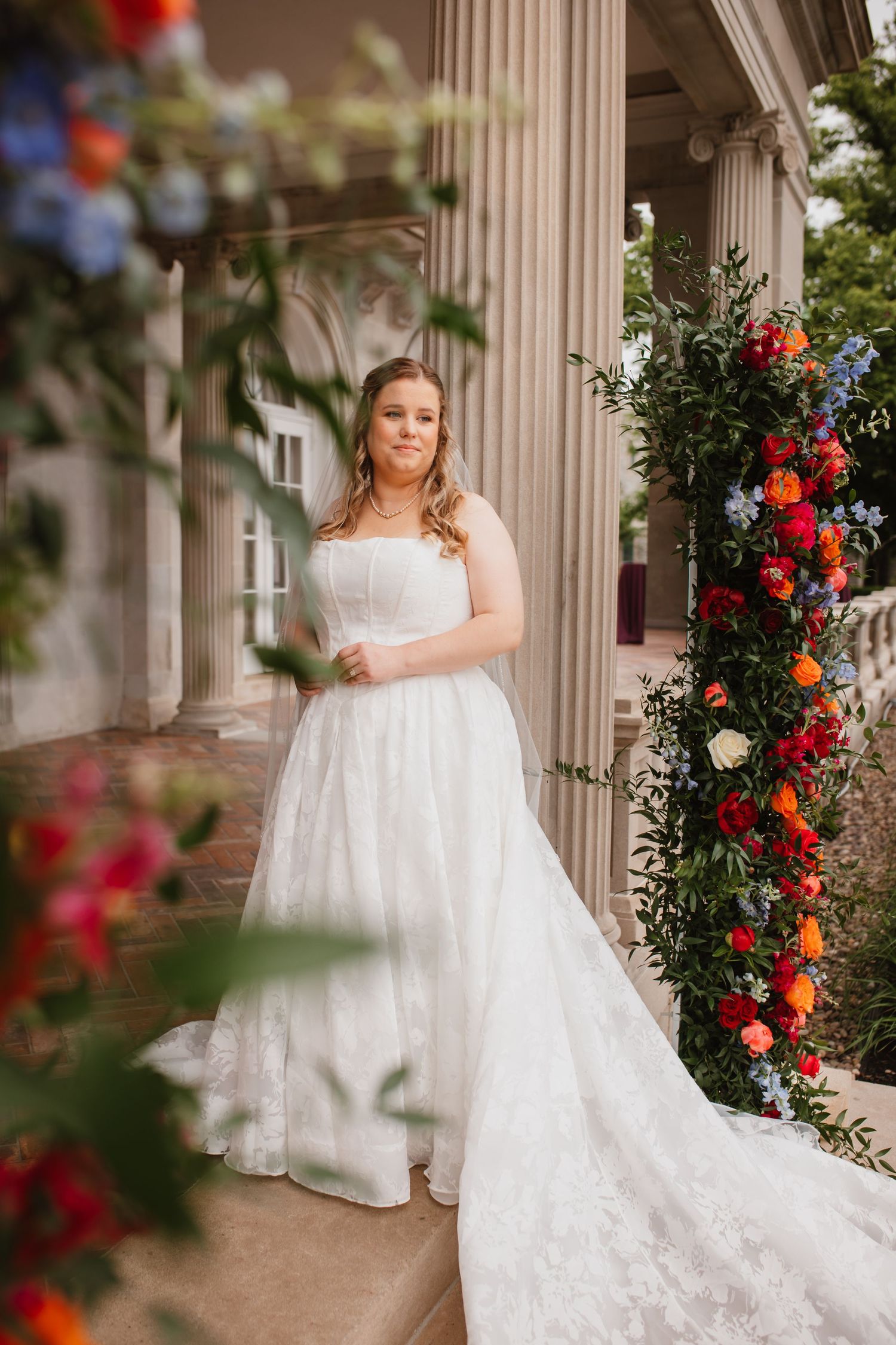 Bride in strapless lace ball gown stands beside vibrant floral arch with colorful roses and columns.