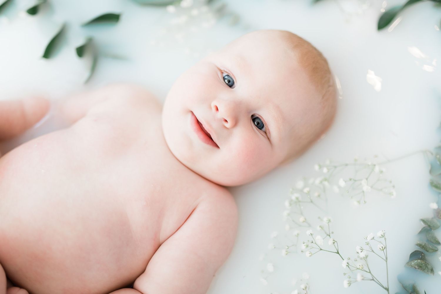 Adorable smiling baby with rosy cheeks lying in milk bath surrounded by white flowers and green leaves.