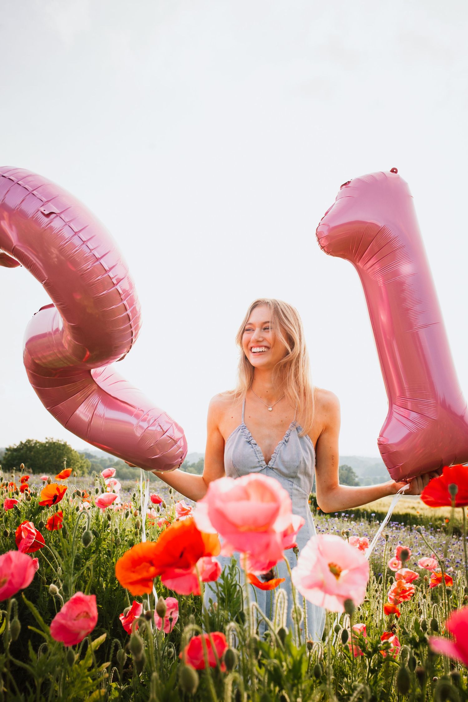 Happy blonde woman celebrating 21st birthday with pink balloons in a colorful poppy field.