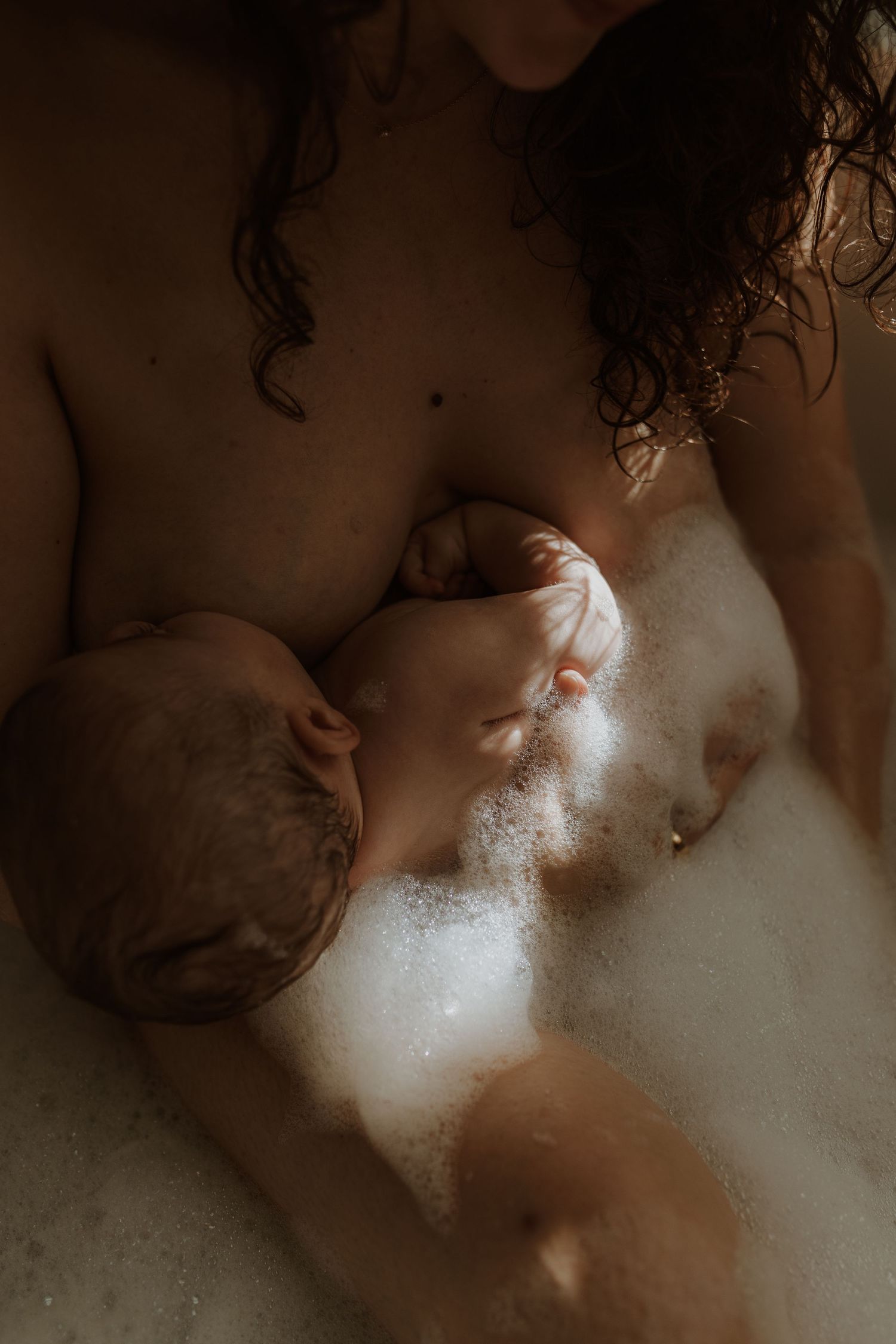 Intimate photo of a mother breastfeeding her newborn in a bubble bath with soft, warm lighting.