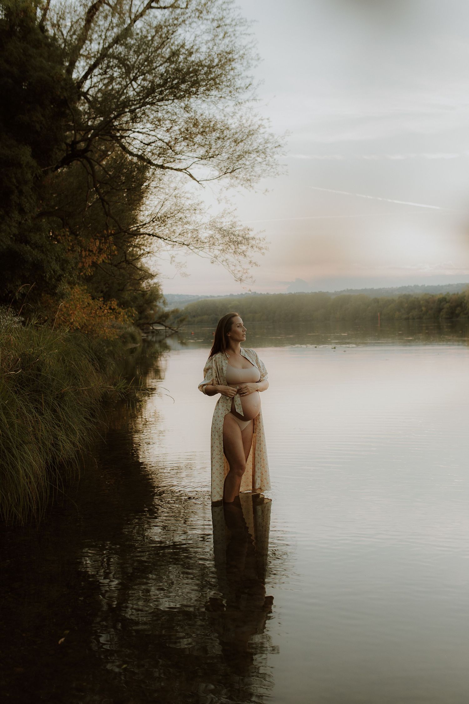 A pregnant woman in a sheer robe stands in calm lake water at sunset, cradling her belly beside a tree-lined shore.