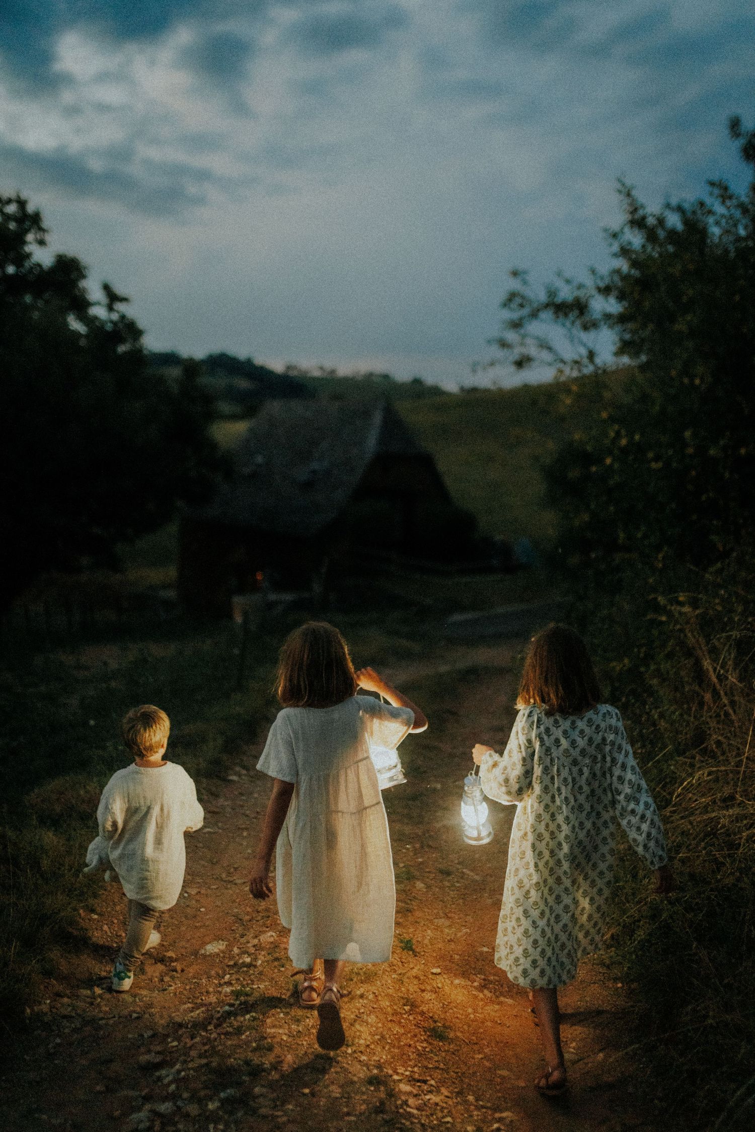 Three children in light summer clothes walk down a dirt path at dusk carrying glowing lanterns toward a rustic cabin.