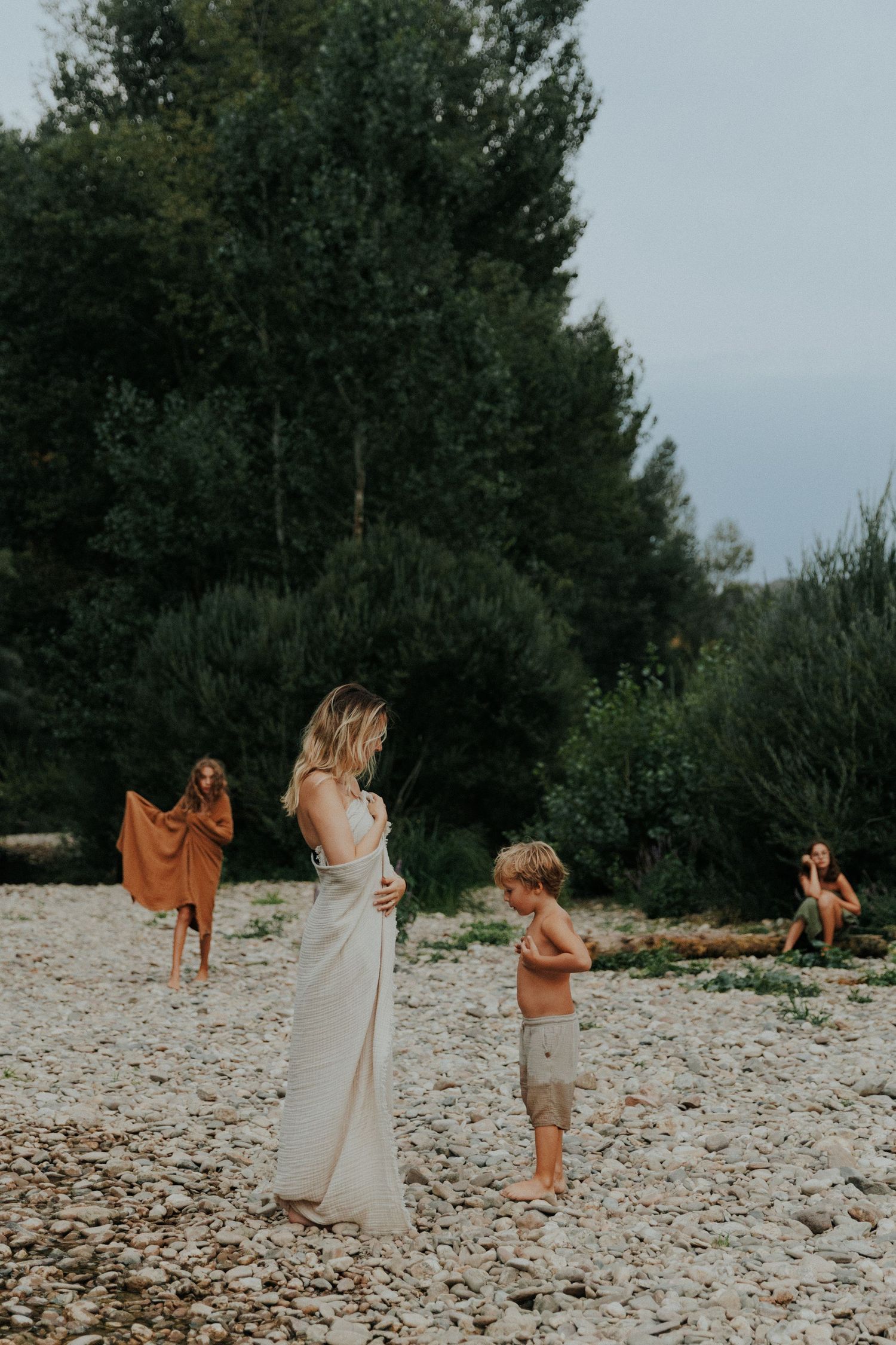Mother in a long white dress stands on a pebble riverbank with her children playing nearby among tall green trees.
