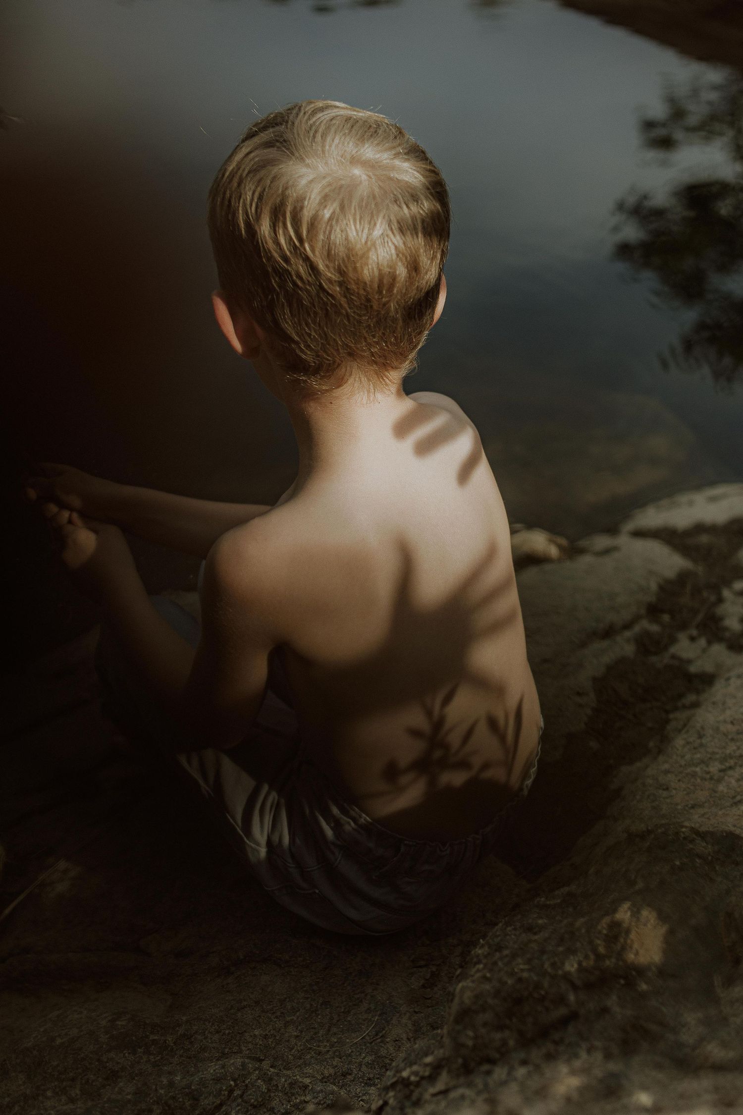 A shirtless blond toddler sits on a rocky shore with dappled shadows on his back, gazing out over still, dark water.