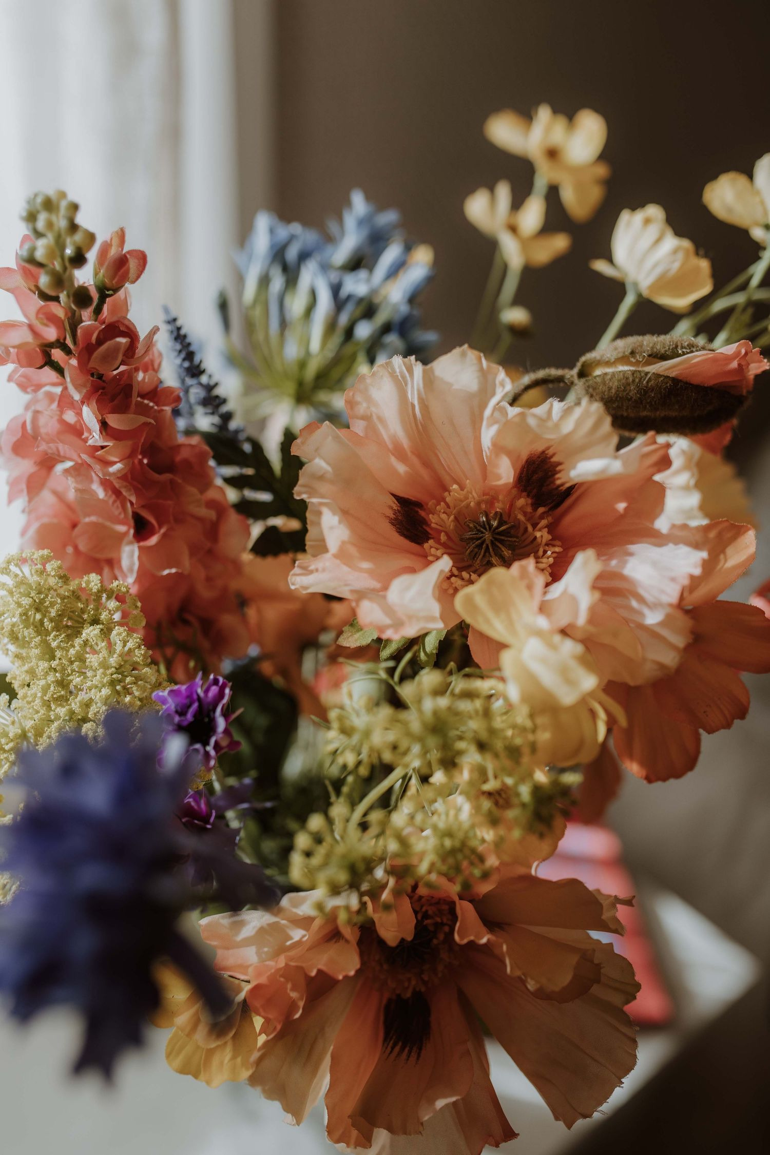 Close-up of a lush bouquet of peach poppies, blue delphinium, and soft yellow wildflowers in natural light.