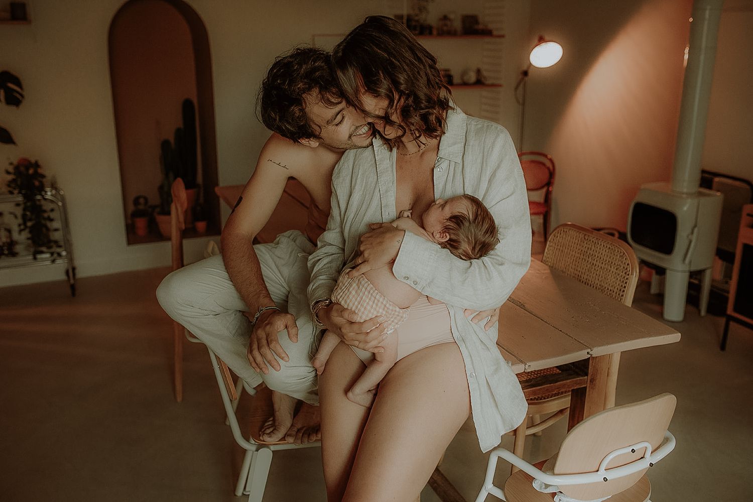 Warm lifestyle family photo of a father embracing a mother who breastfeeds their baby in a cozy home interior.